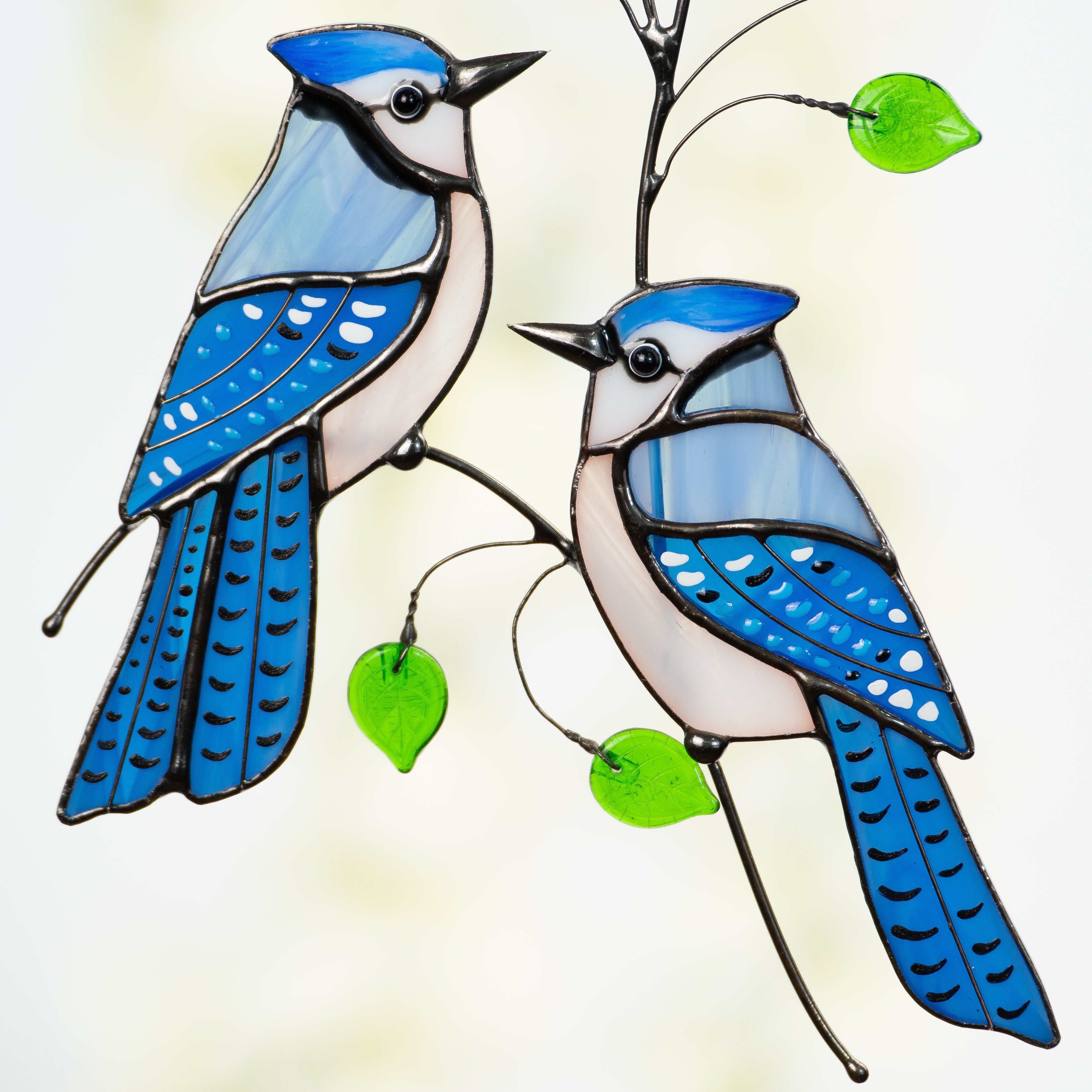 Zoomed view of two blue stained glass jays perched on a curved branch with green leaves and vivid blue feathers