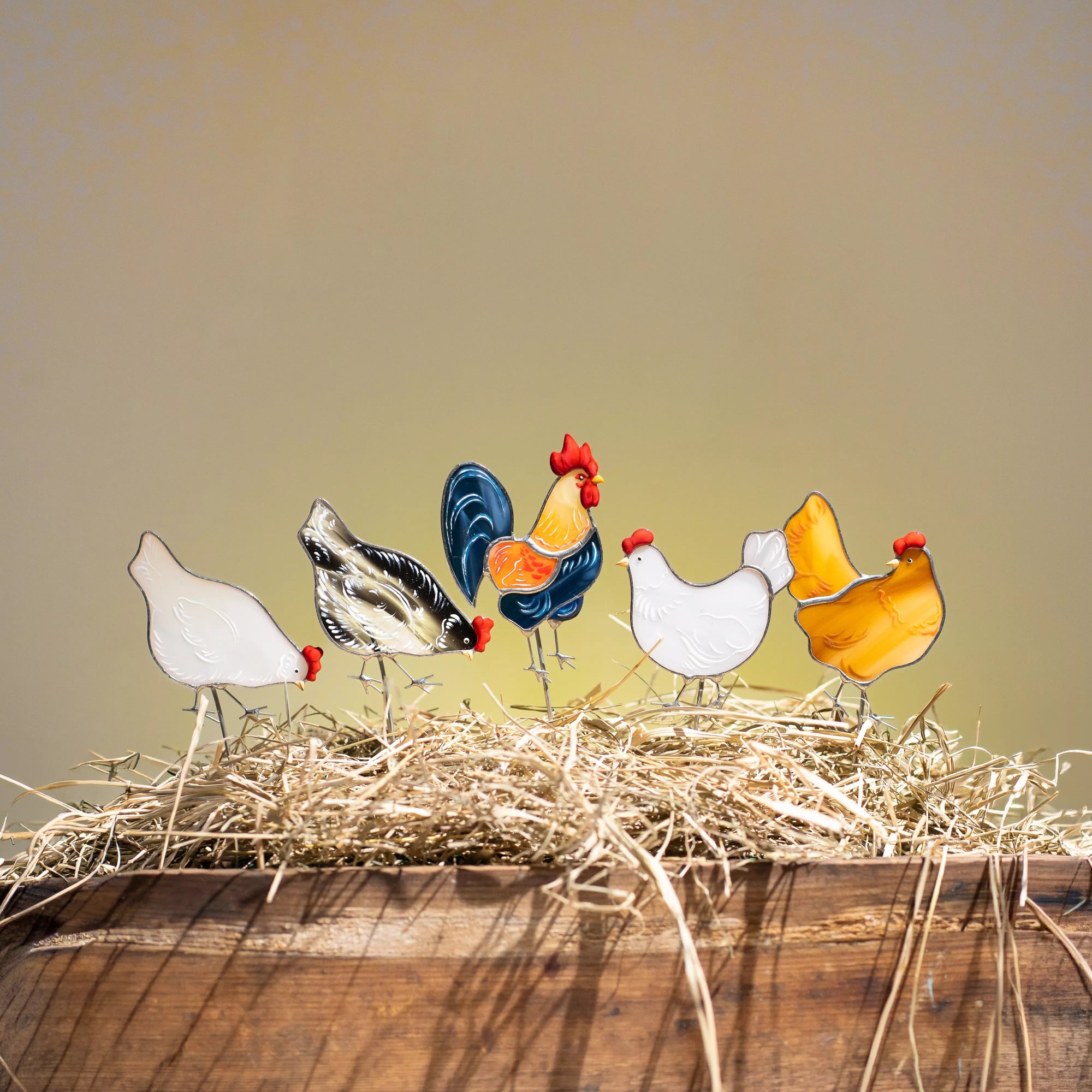 Colorful glass chicken figurines on a bed of hay with a wooden surface and beige background