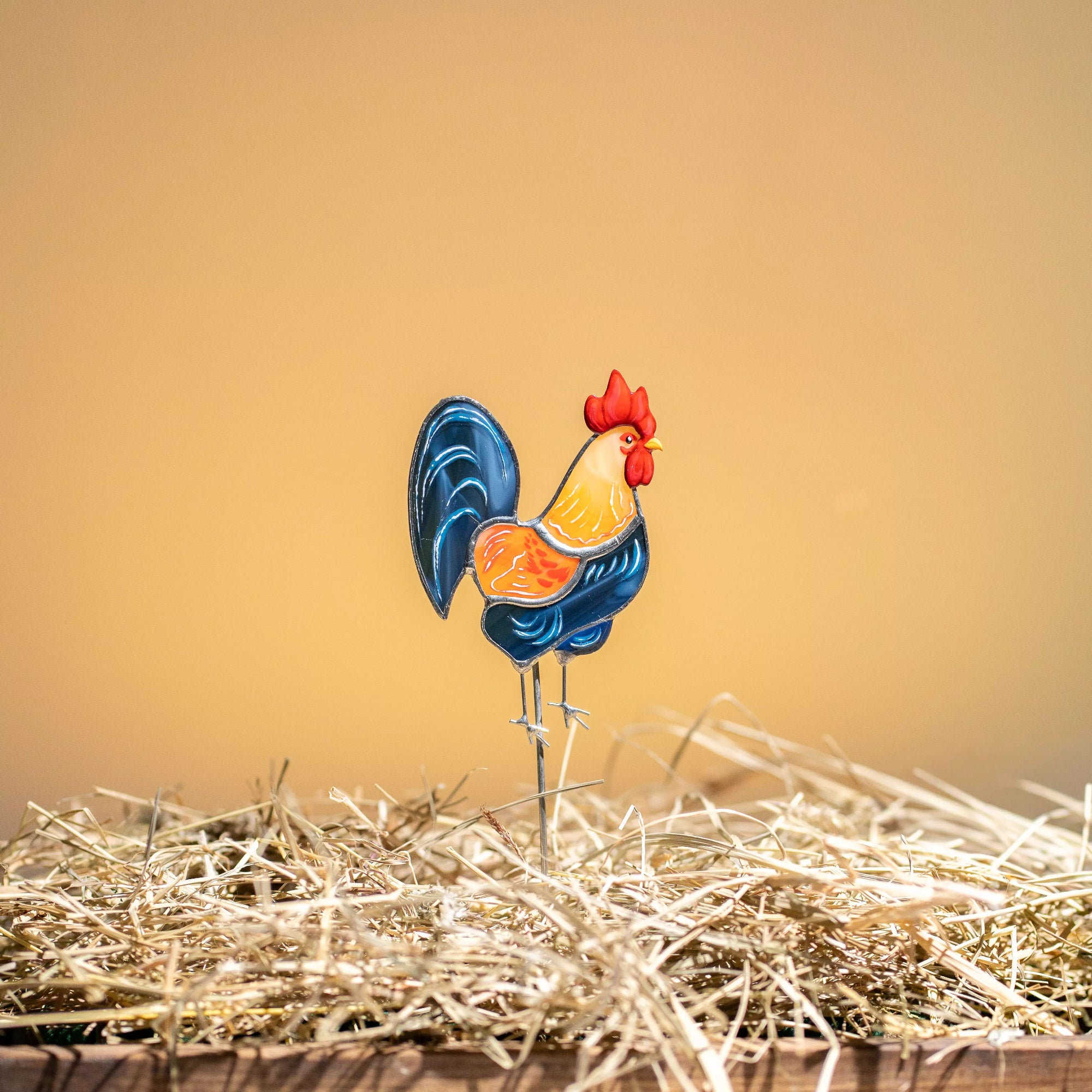 Colorful stained glass rooster plant stake on a bed of straw with a warm background