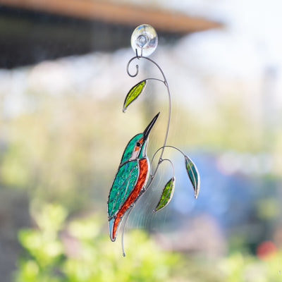 Kingfisher stained glass suncatcher hanging on a window with a suction cup, blurred garden background.