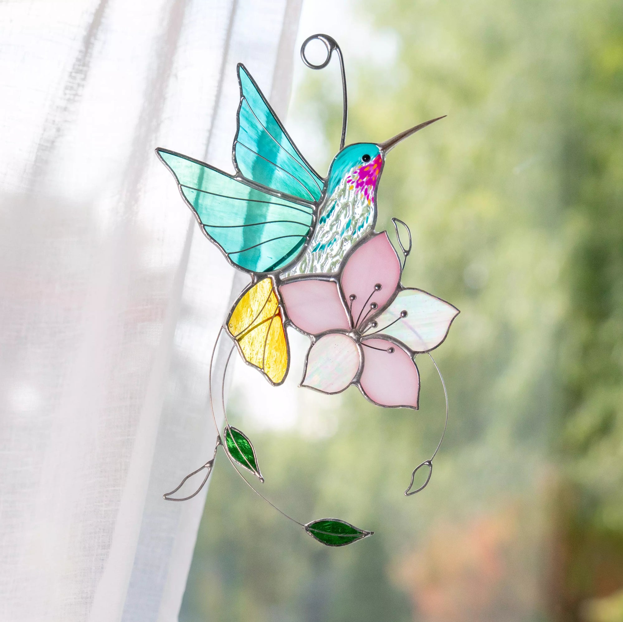 Stained glass hummingbird and flower decoration against a blurred green outdoor background.