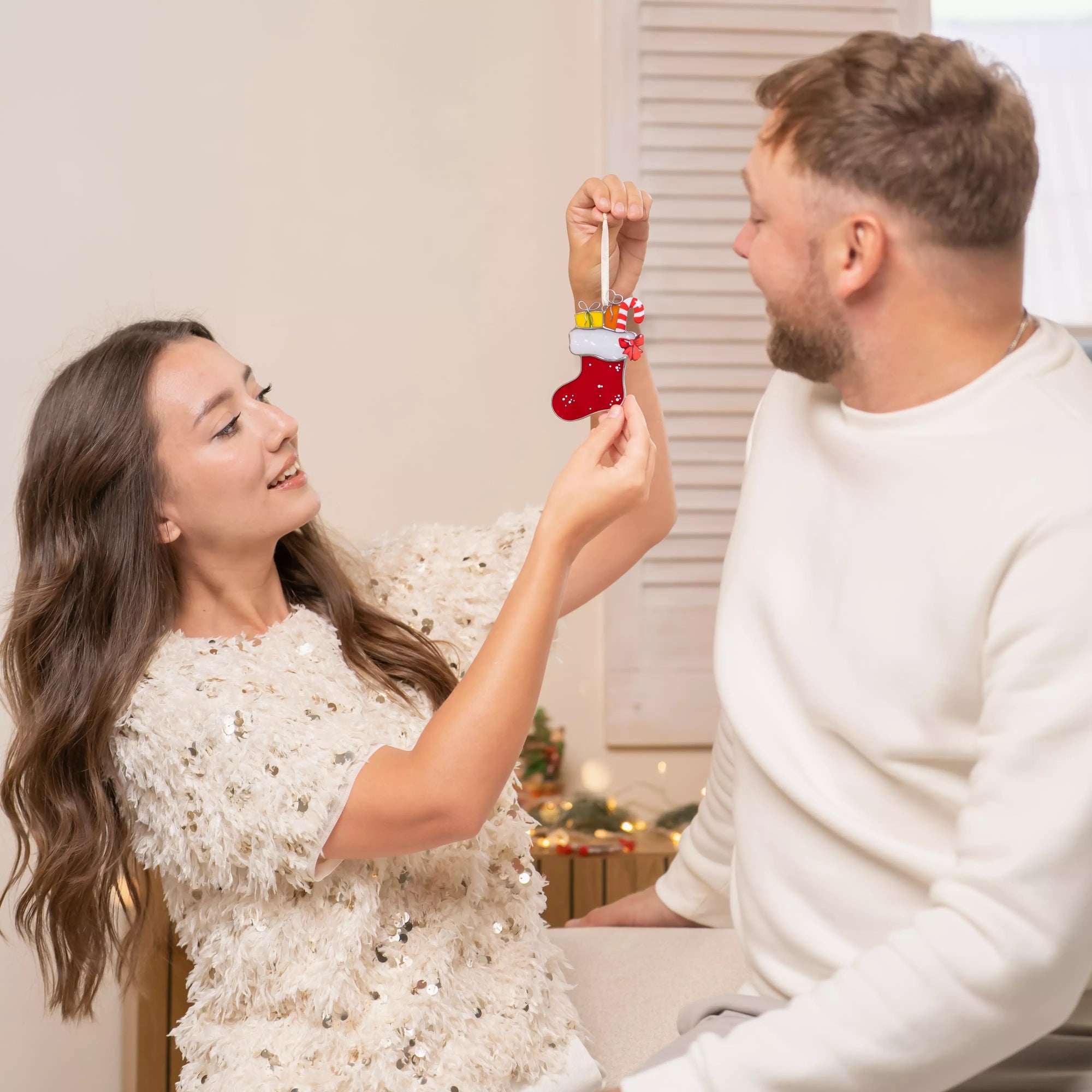 Smiling couple looking at a stained glass red Christmas stocking ornament with gifts and candy cane in cozy festive room.