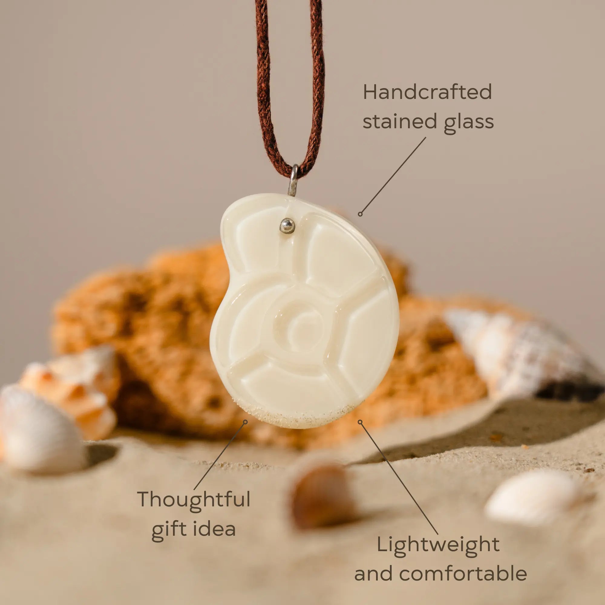 Cream-colored stained glass nautilus pendant hanging above sand with shells. Labeled as a Thoughtful gift idea  and Lightweight and comfortable handcrafted pendant