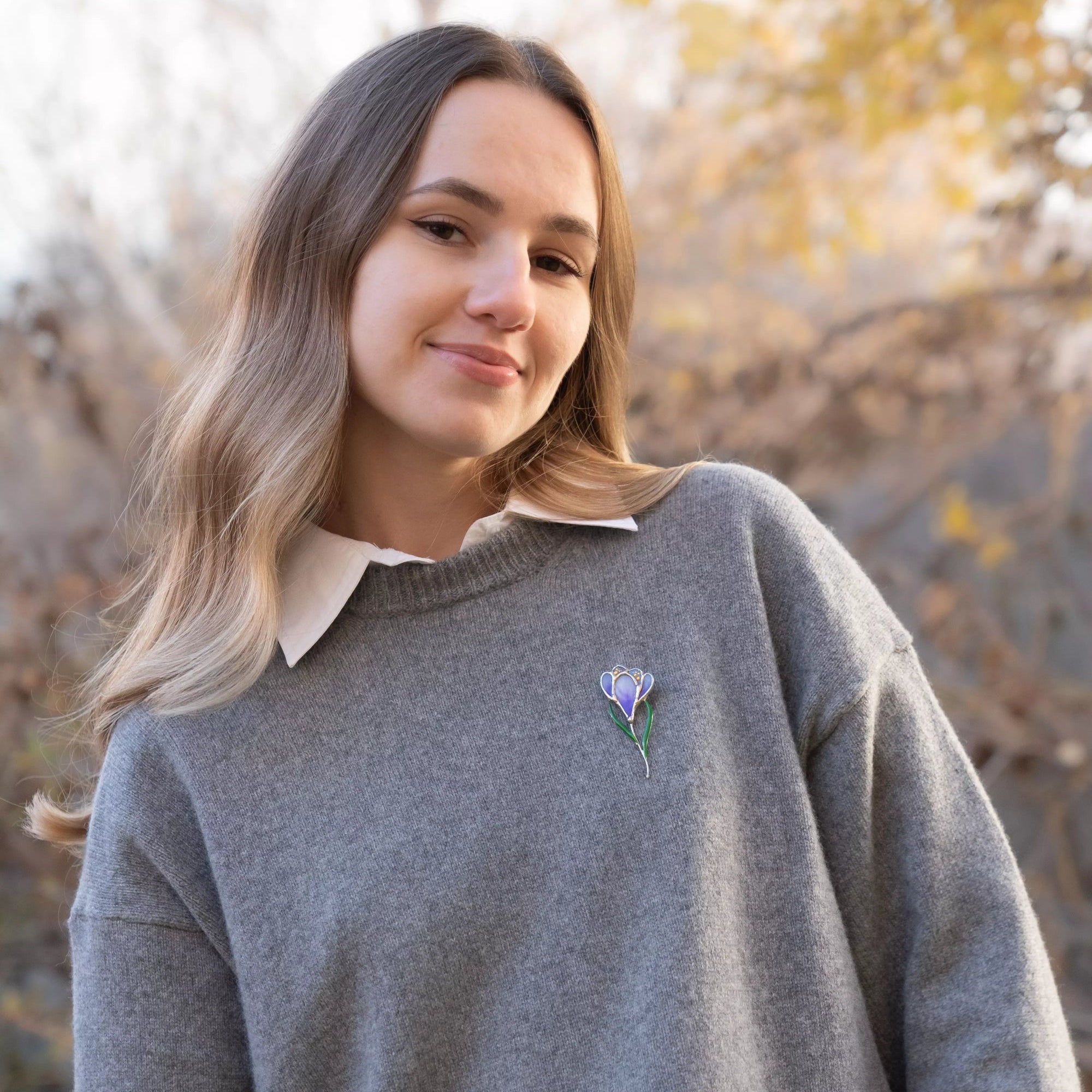 Woman wearing a purple stained glass crocus brooch pinned to a gray sweater, showing scale and everyday styling.