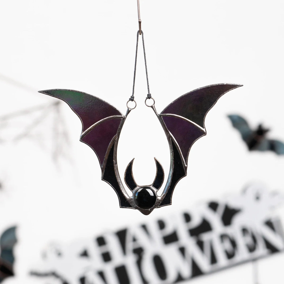 Iridescent bat stained glass suncatcher hanging on a cord in front of a Happy Halloween sign and smaller glass bats - handmade gothic window decor for the season