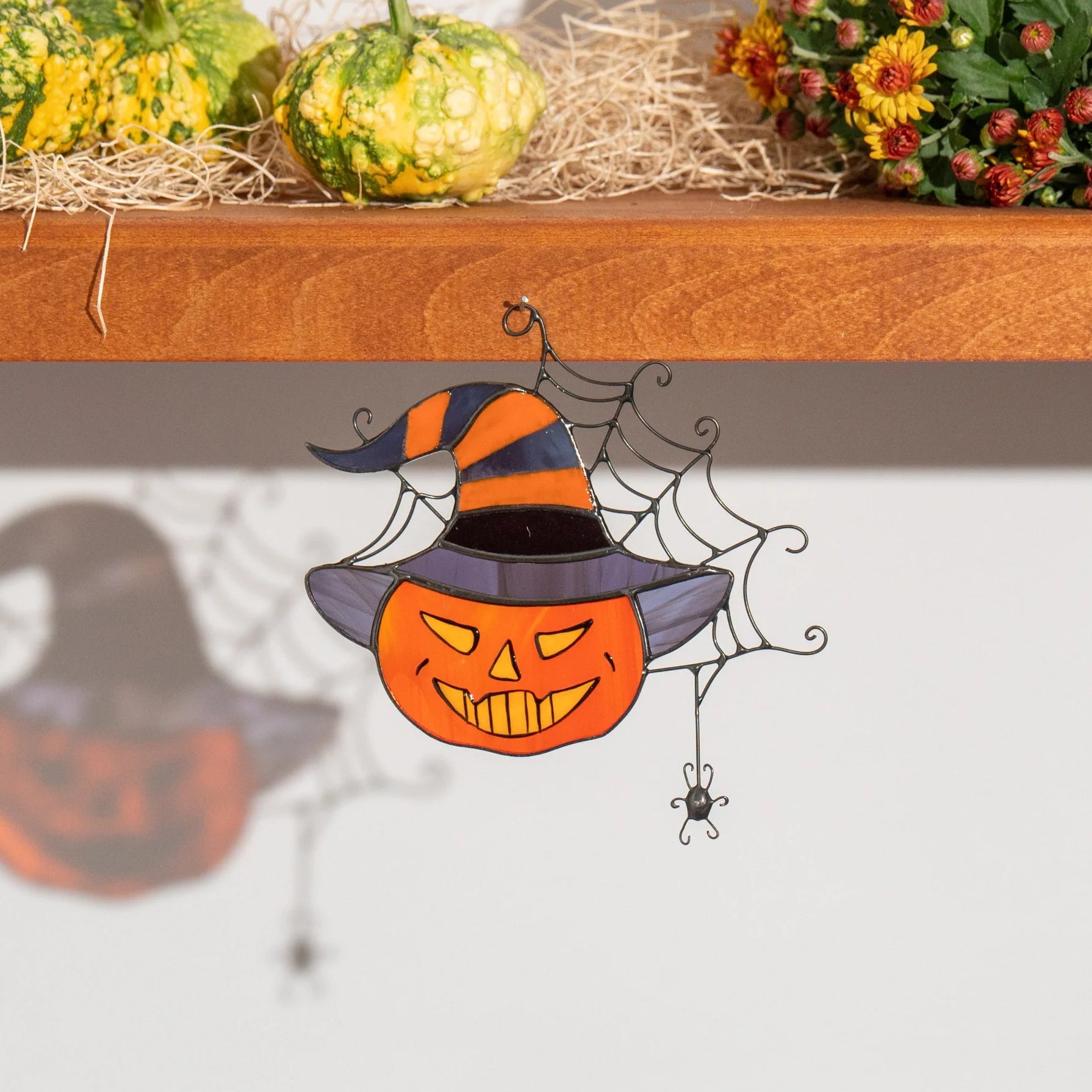 Stained glass pumpkin-in-a-witch-hat suncatcher hanging below a shelf of gourds, hay and autumn mums, casting a spider-web shadow on the wall - grinning jack-o'-lantern Halloween window decor