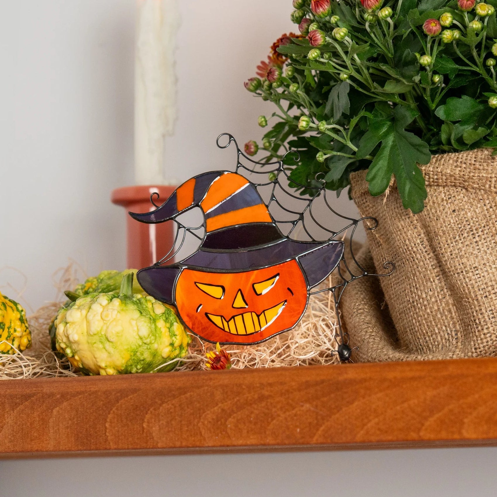 Stained glass jack-o'-lantern in a witch hat suncatcher on a fall shelf beside a burlap pot of mums, a candle and gourds - handmade grinning pumpkin with a wire spider web, Halloween decor