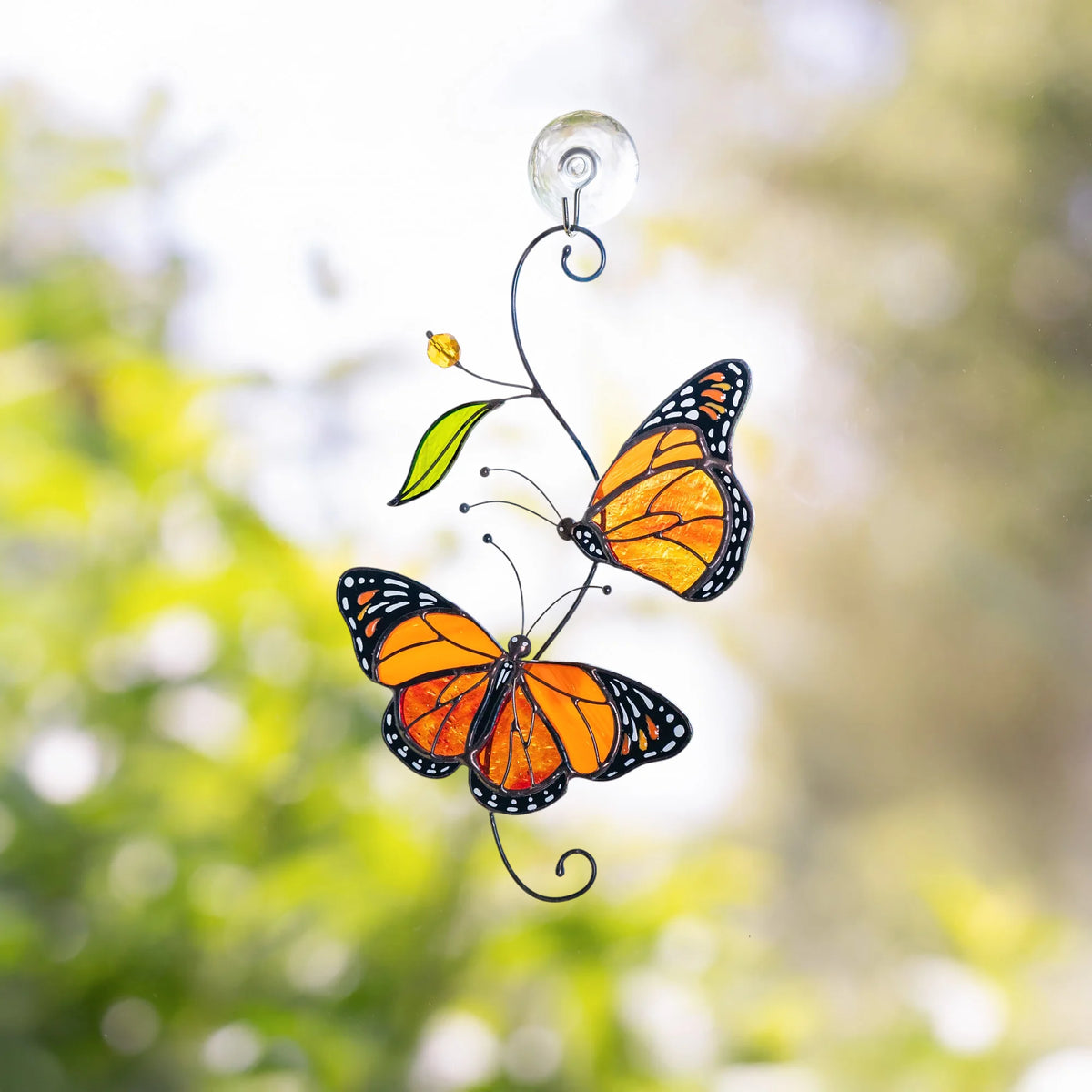 Pair of monarch butterflies stained glass suncatcher hanging on a clear suction cup at a sunny window, green garden bokeh behind - handmade orange glass art for bright rooms