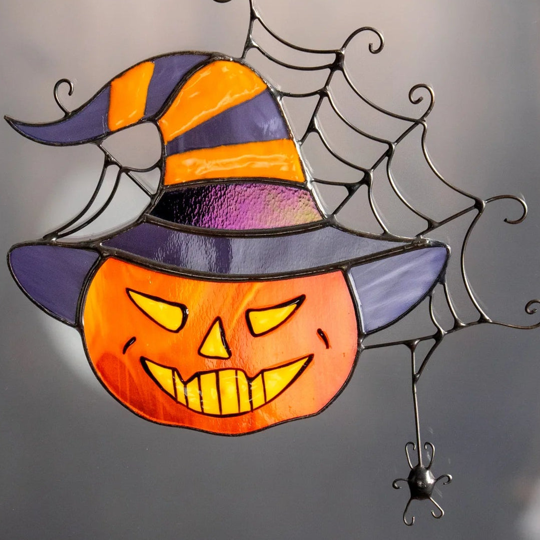 Grinning stained glass pumpkin in a witch hat suncatcher stuck to a window with a suction cup, a ghostly Halloween scene beyond - handmade jack-o'-lantern with a wire spider web and dangling spider