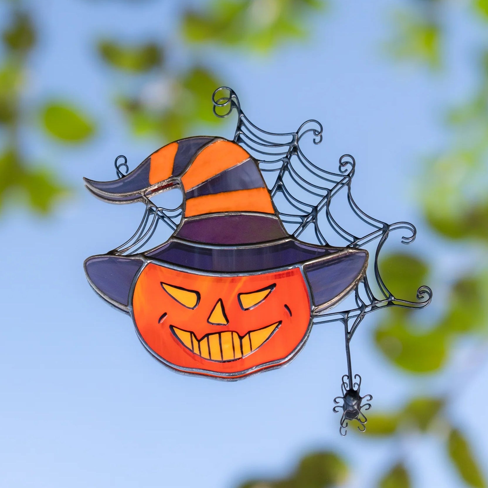 Stained glass pumpkin in a witch hat suncatcher against a blue sky and green leaves, orange jack-o'-lantern face glowing in sunlight with a wire spider web and spider - handmade Halloween window decor