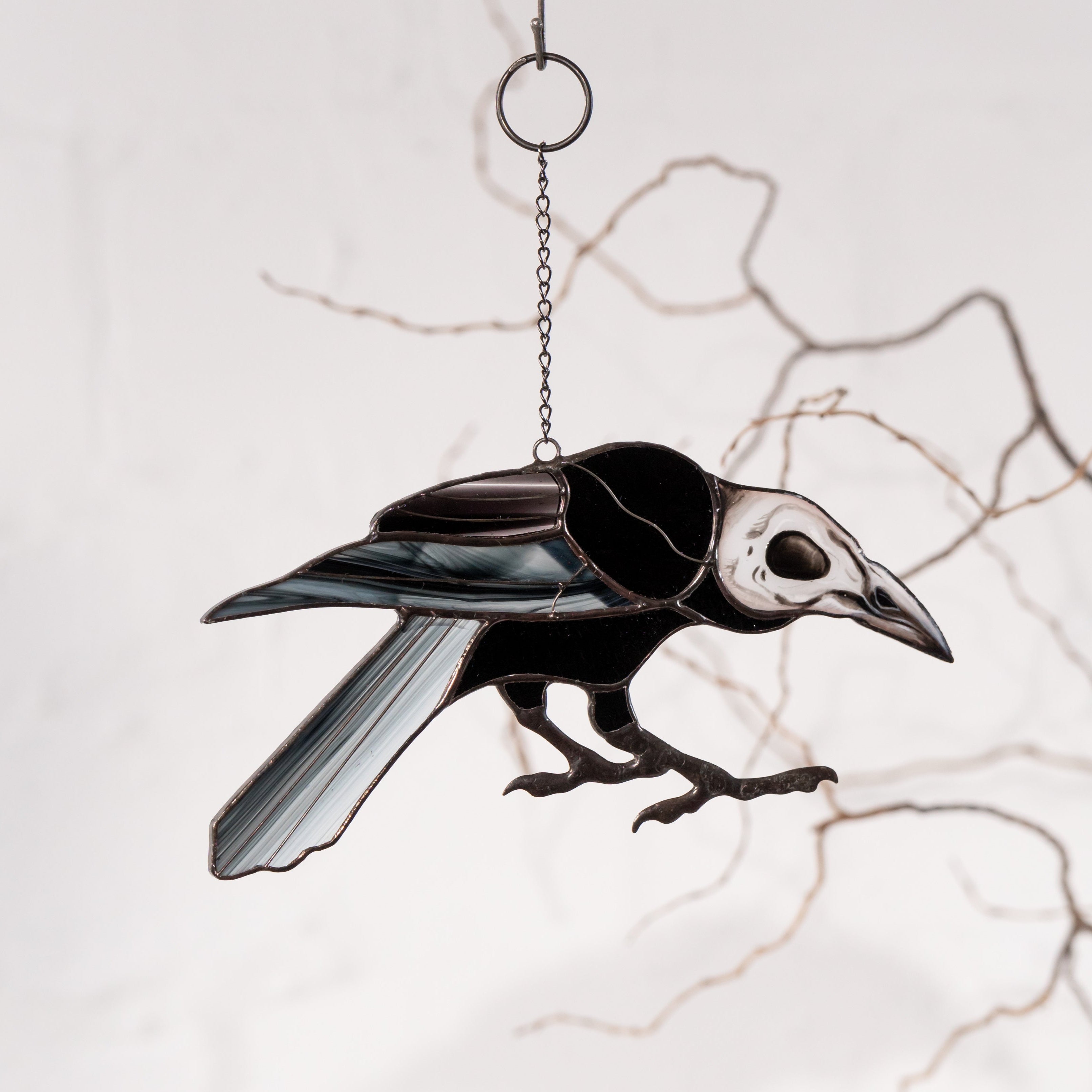 Raven suncatcher with skull face hanging against a pale wall and artistic dry branches in the background.