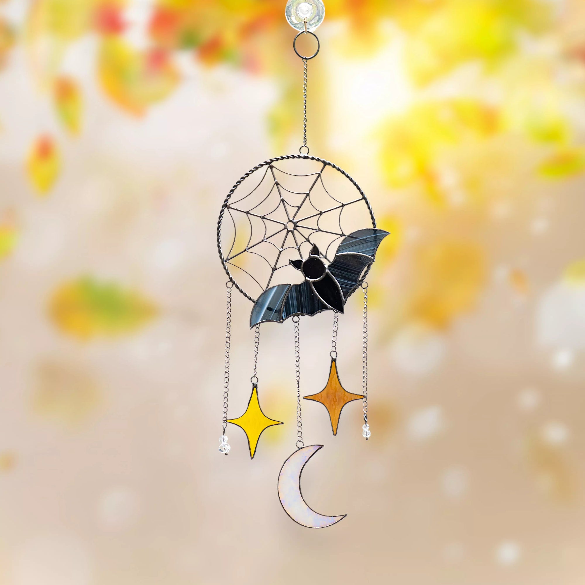 Decorative stained glass Halloween ornament with bat, spider web, stars, and moon against a blurred autumn background.
