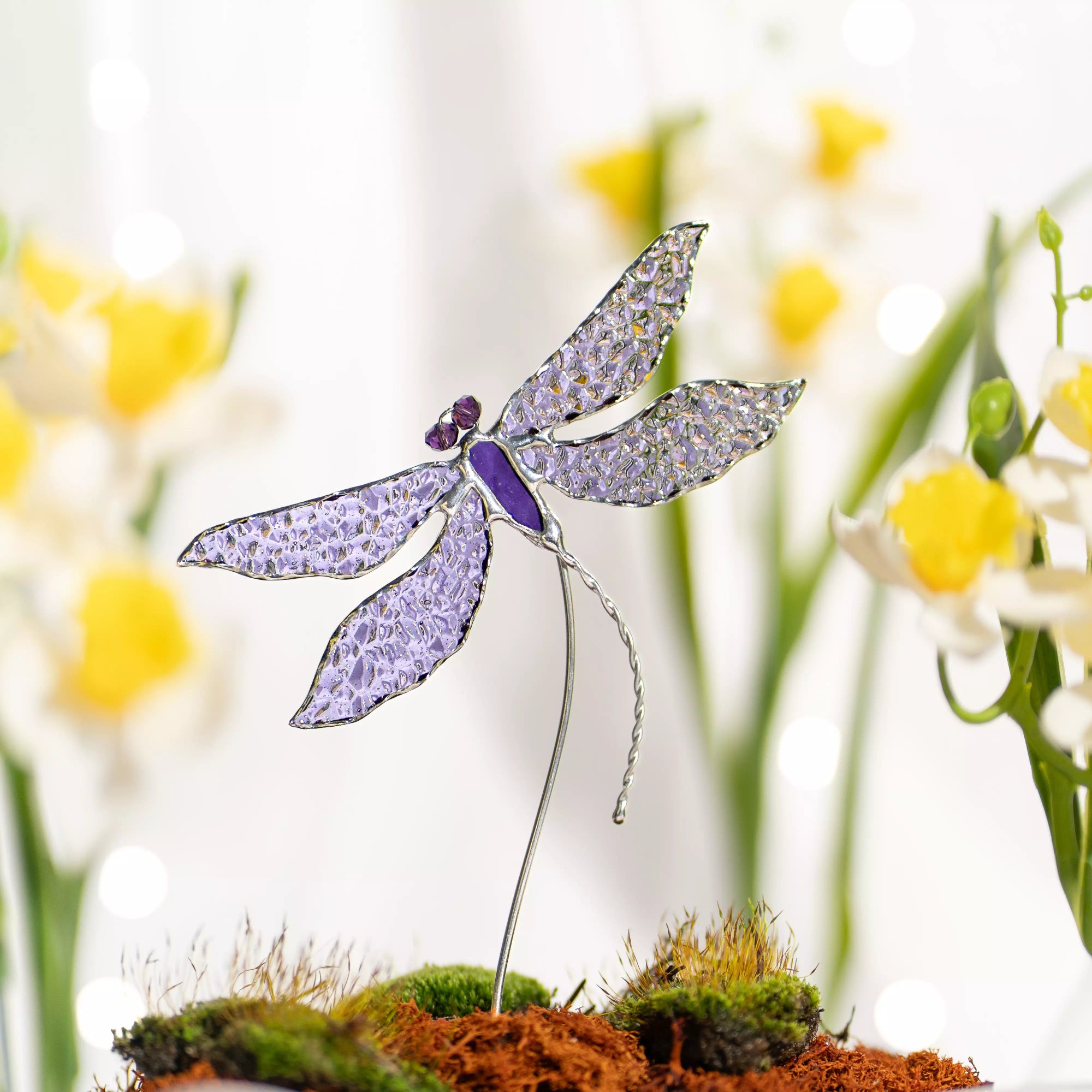 Stained glass dragonfly plant stake on a stand with a blurred floral background