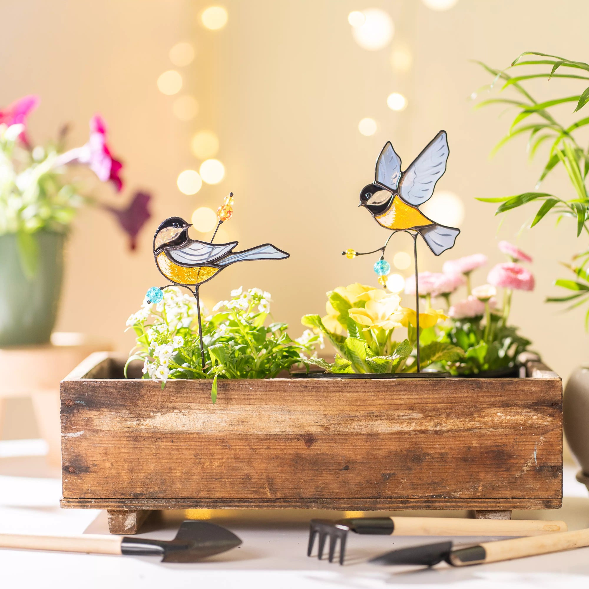 Stained glass garden stakes in wooden planter, flowers, and bird figurines.