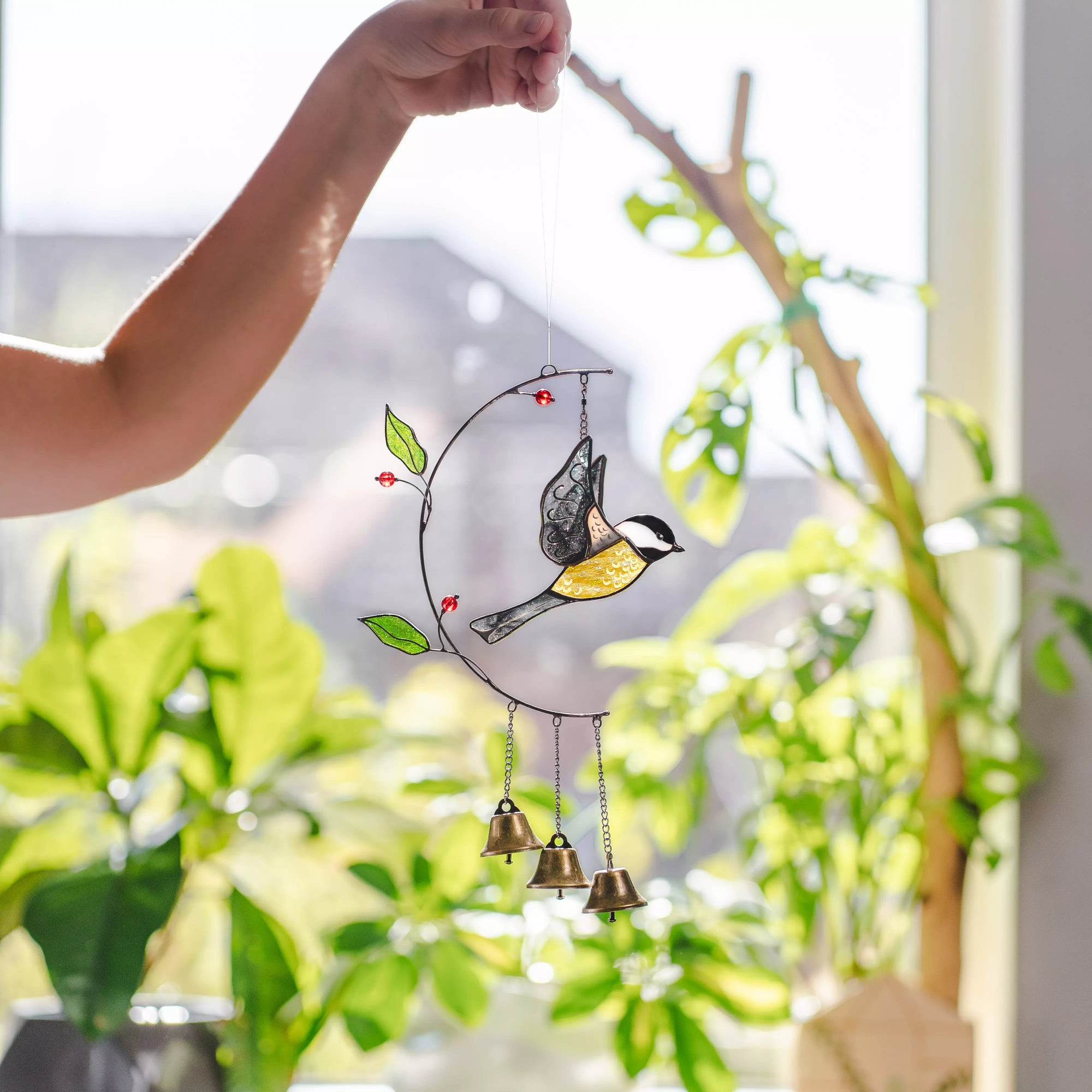 Stained glass chickadee ornament with bells hanging in front of a window with plants.