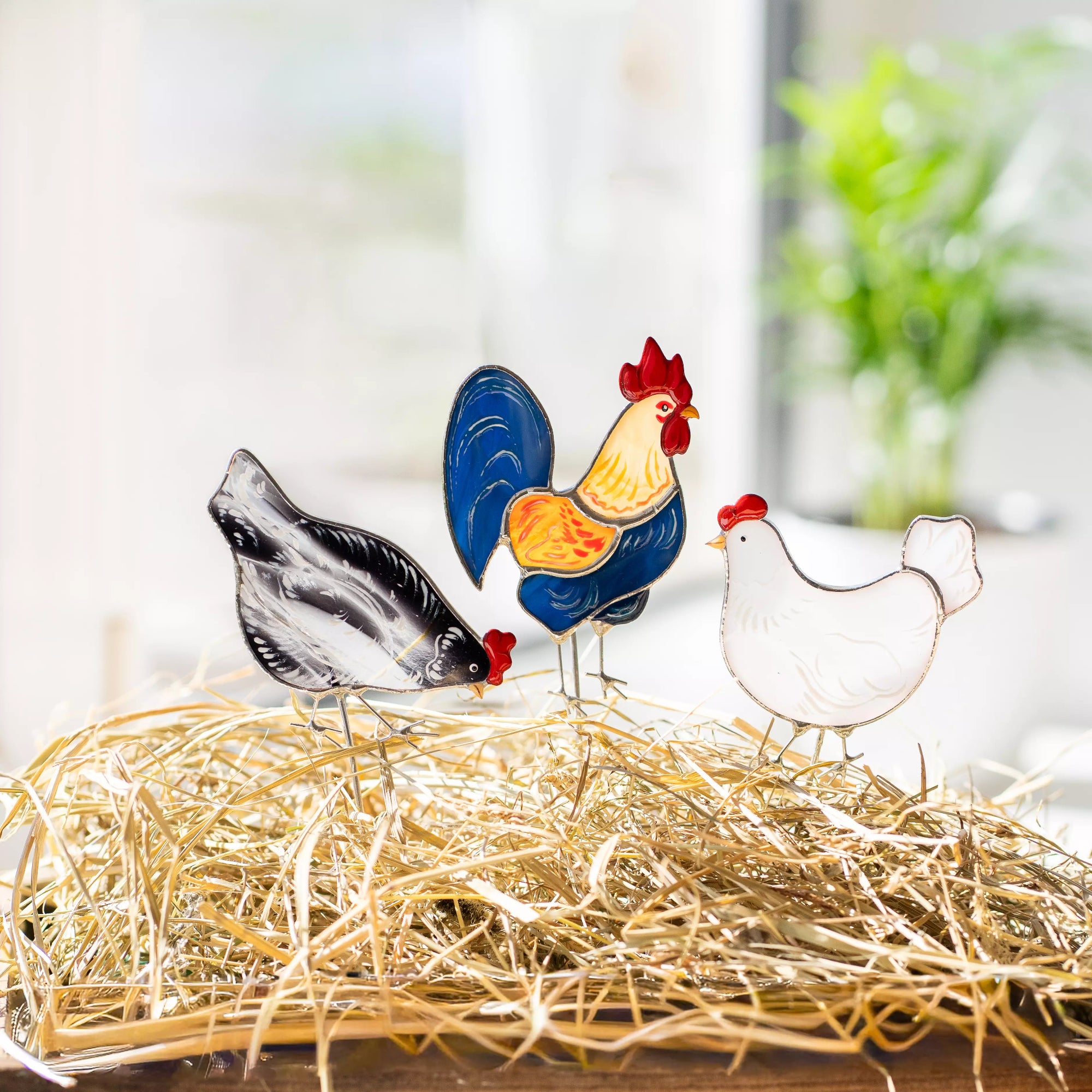 Stained glass rooster and two chickens on a bed of straw with a blurred indoor background.