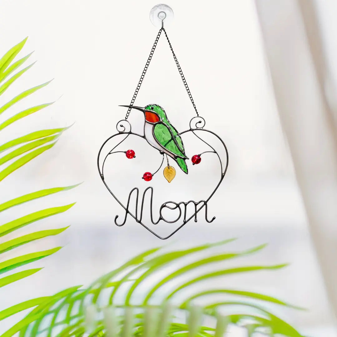 Stained glass hanging heart with a hummingbird and 'Mom' text, surrounded by green leaves.