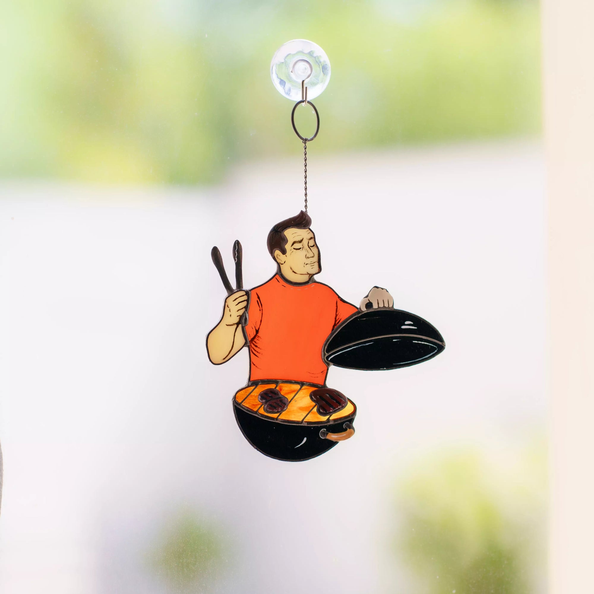 Window hanging stained glass ornament of a chef cooking on a grill with a suction cup on a blurred background