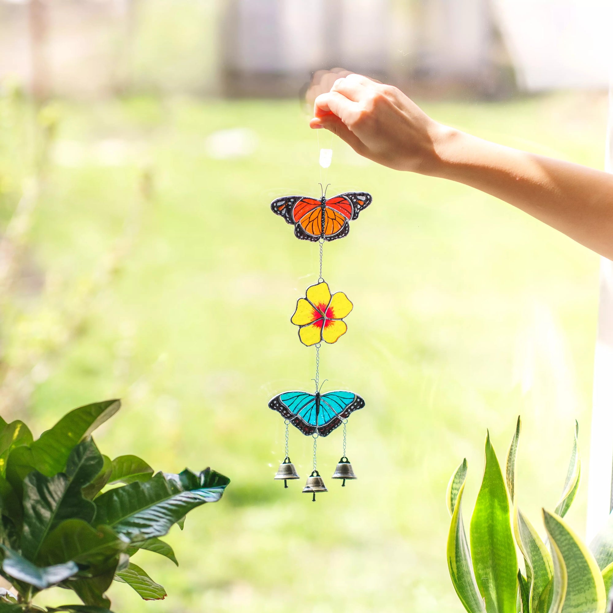 Stained glass wind chime with butterflies and flowers held by a hand against a blurred green outdoor background