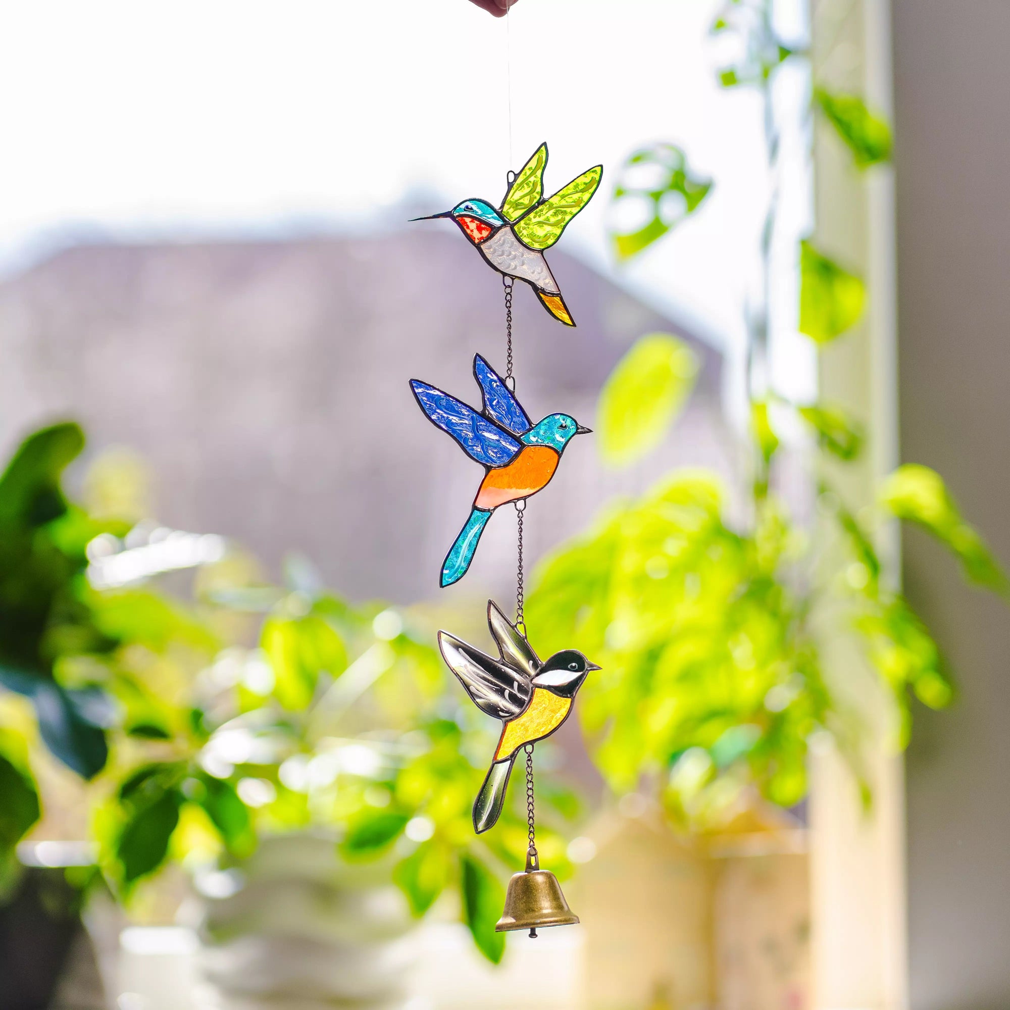 Stained glass hanging with colorful birds and a bell against a blurred greenery background