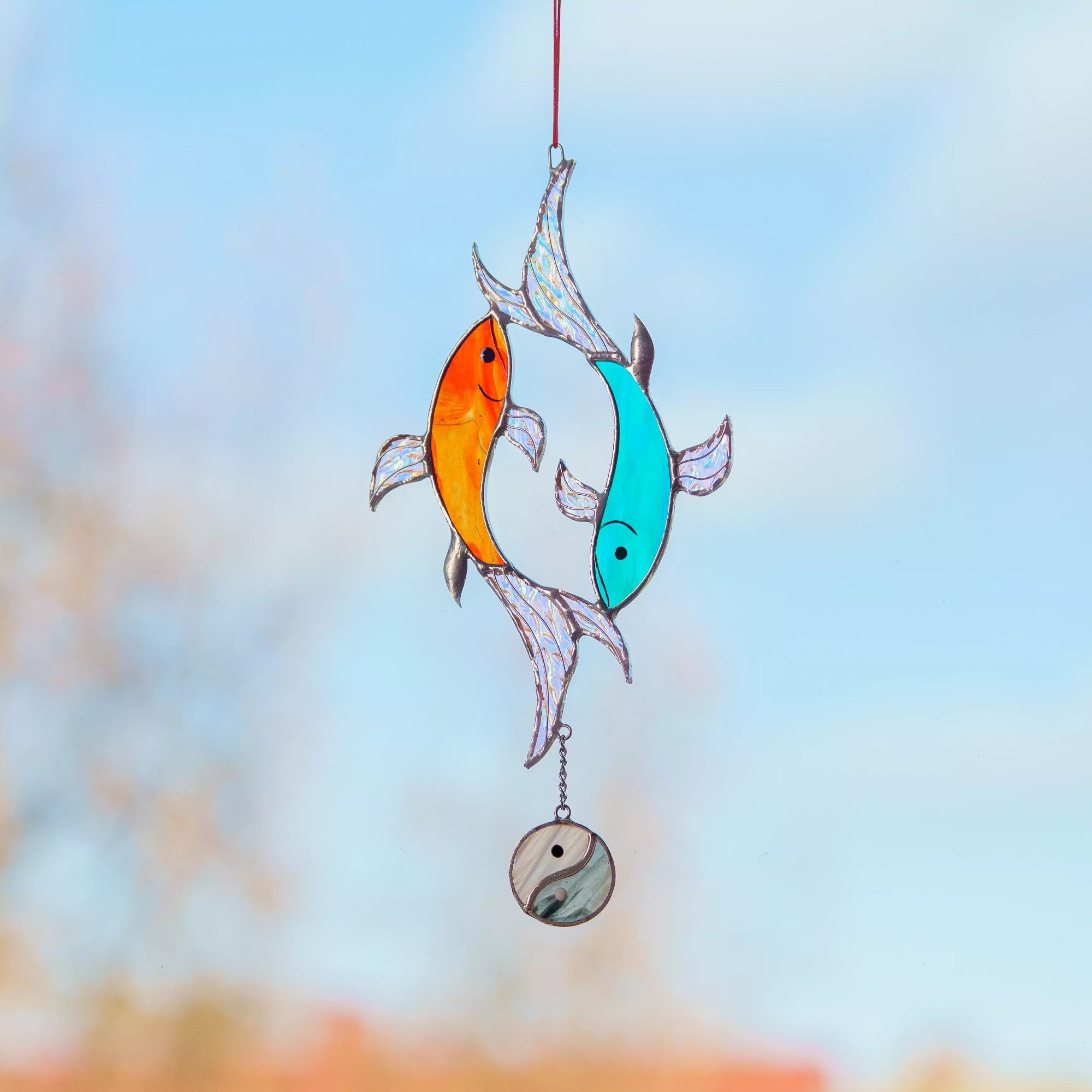 Stained glass window hanging with two fish, one orange and one blue, against a blurred natural background.