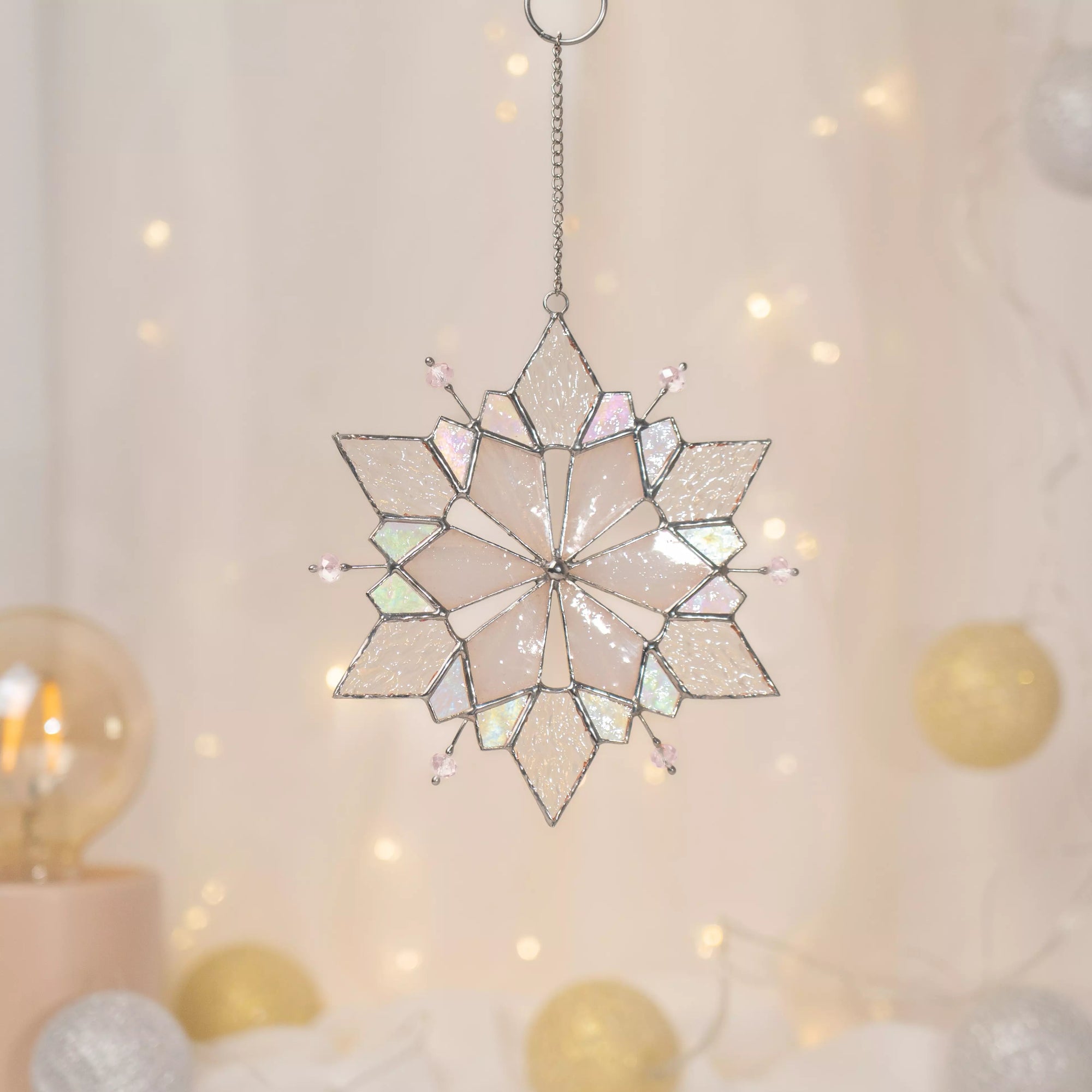 Stained glass snowflake ornament made with iridescent glass hanging against a soft, blurred background with lights.