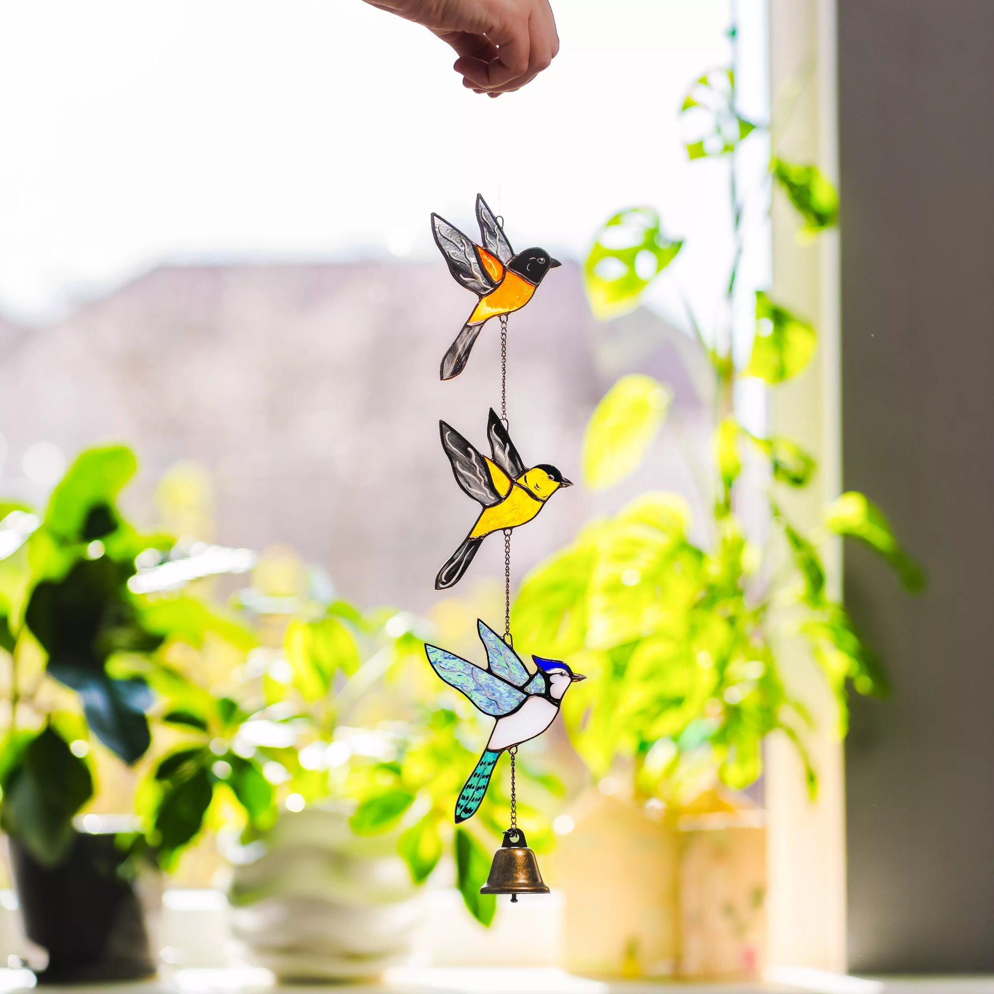 Decorative stained glass birds wind chime with a bell hanging in front of a window with plants.