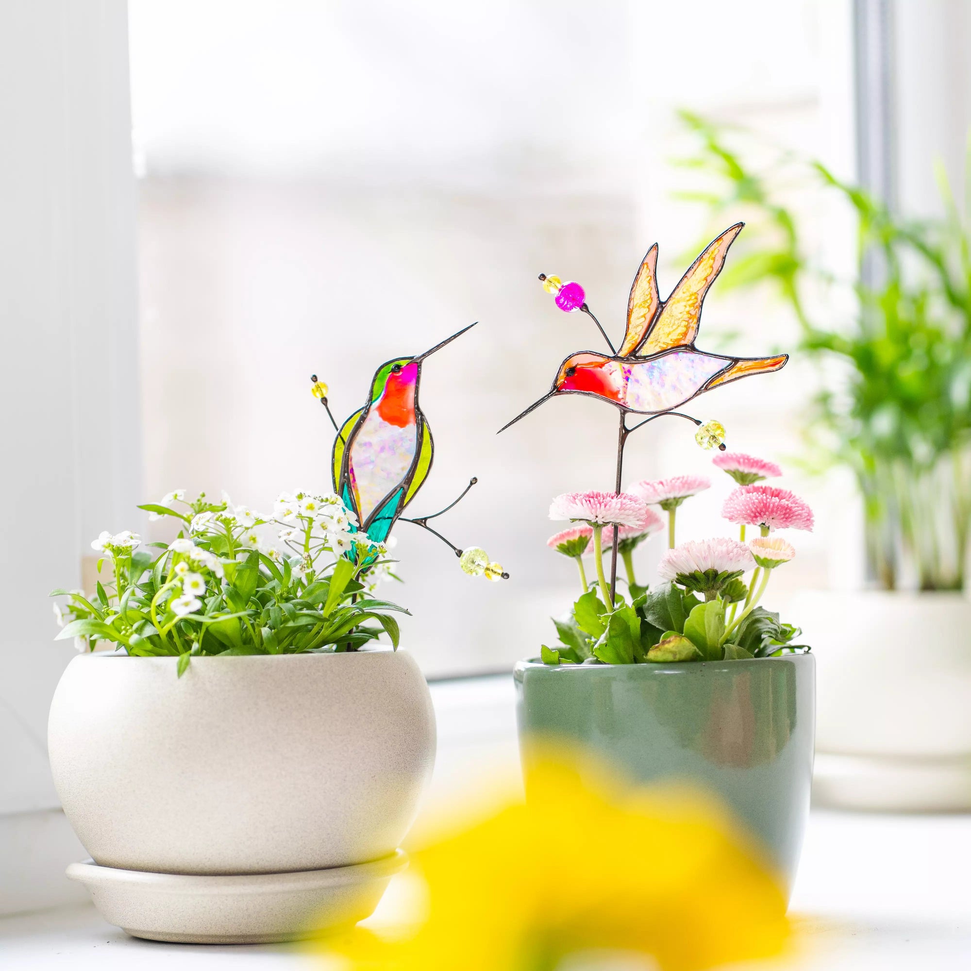 Stained glass hummingbird plant stakes with plants on a windowsill.