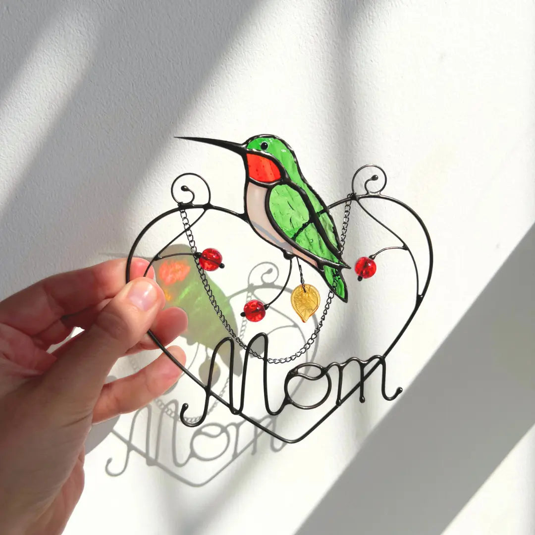 Stained glass suncatcher with a green bird and 'Mom' text, held by a hand.