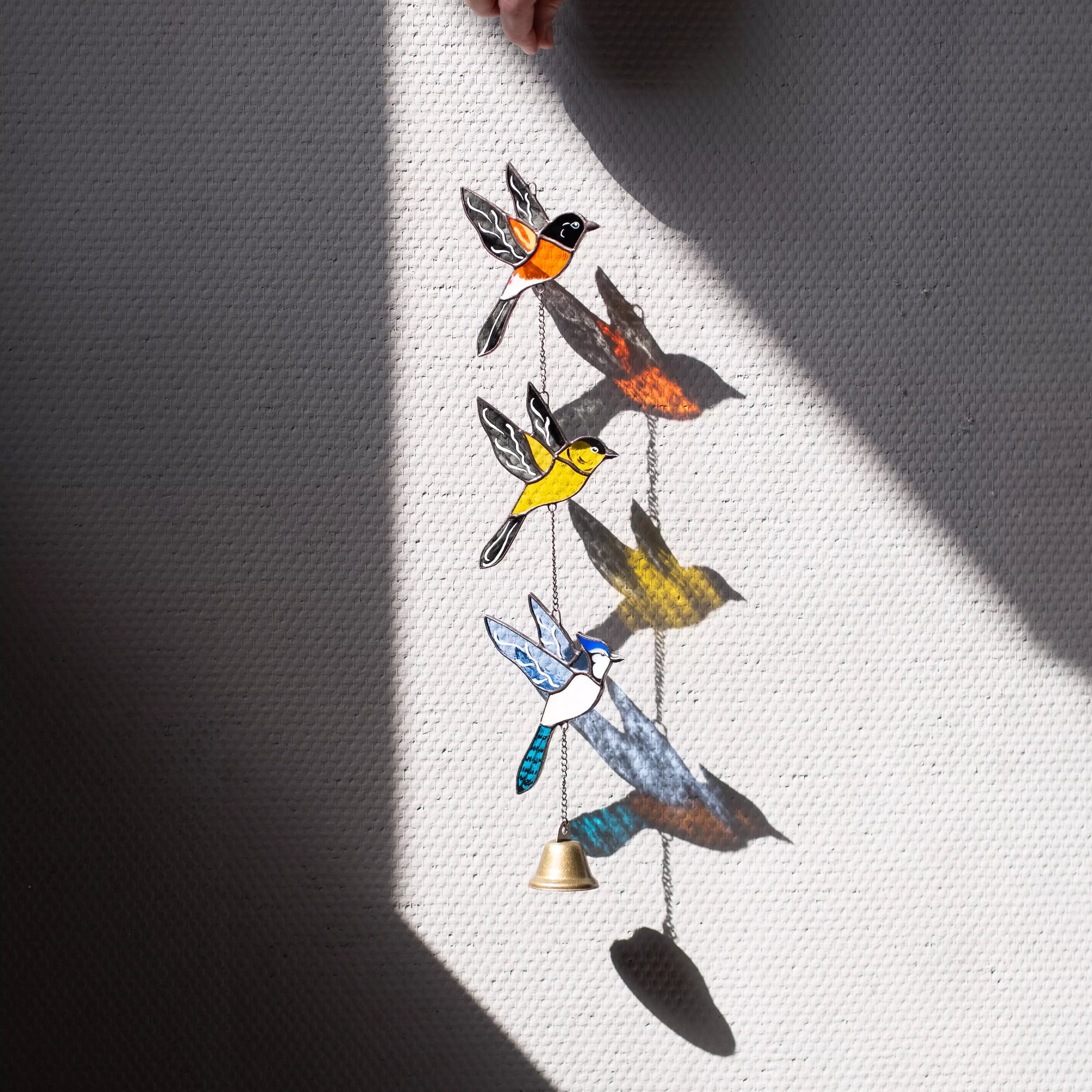 Staiend glass wall hanging with colorful birds and a bell on a textured white surface.