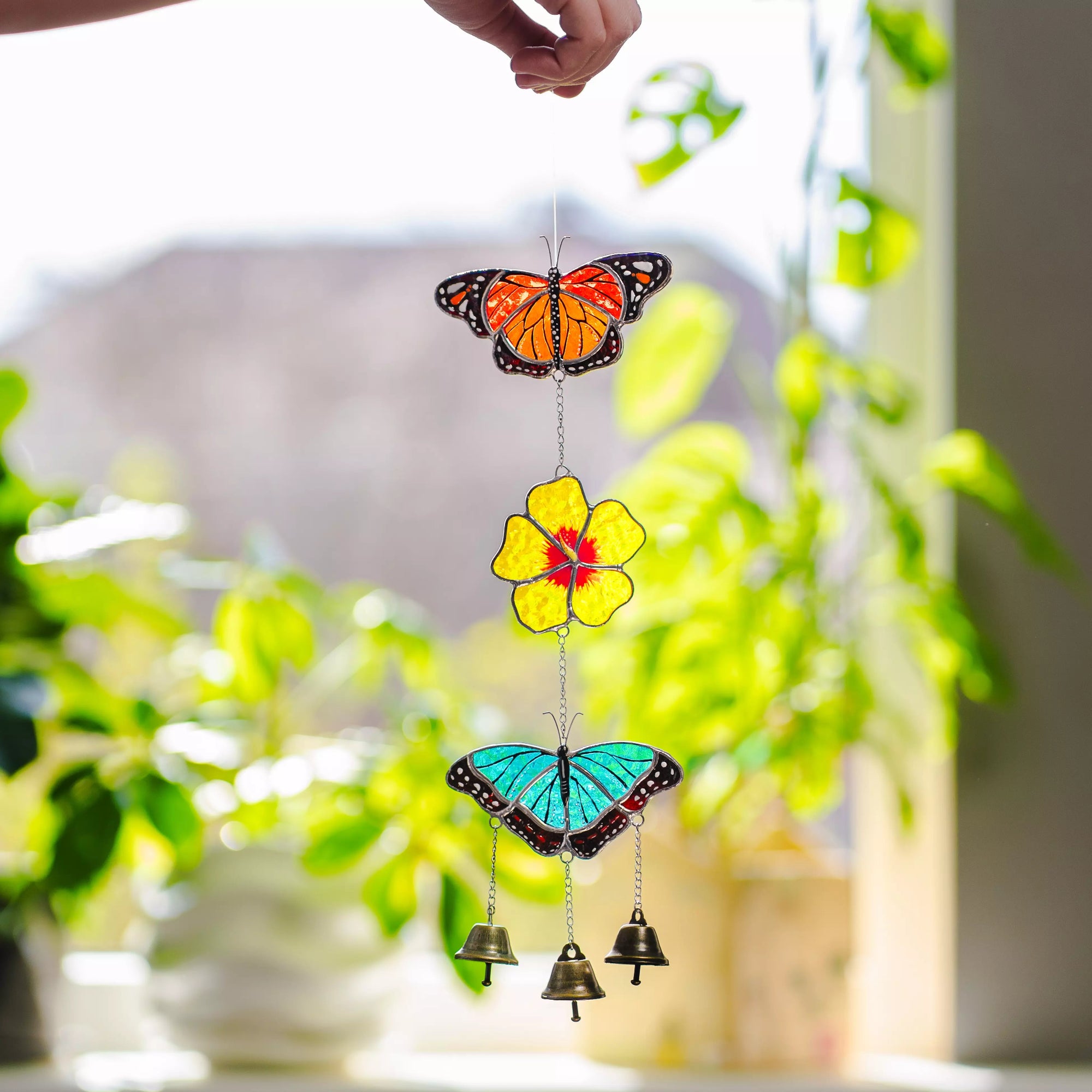 Stained glass wind chime with butterfly and flower design in front of a blurred indoor plant setting