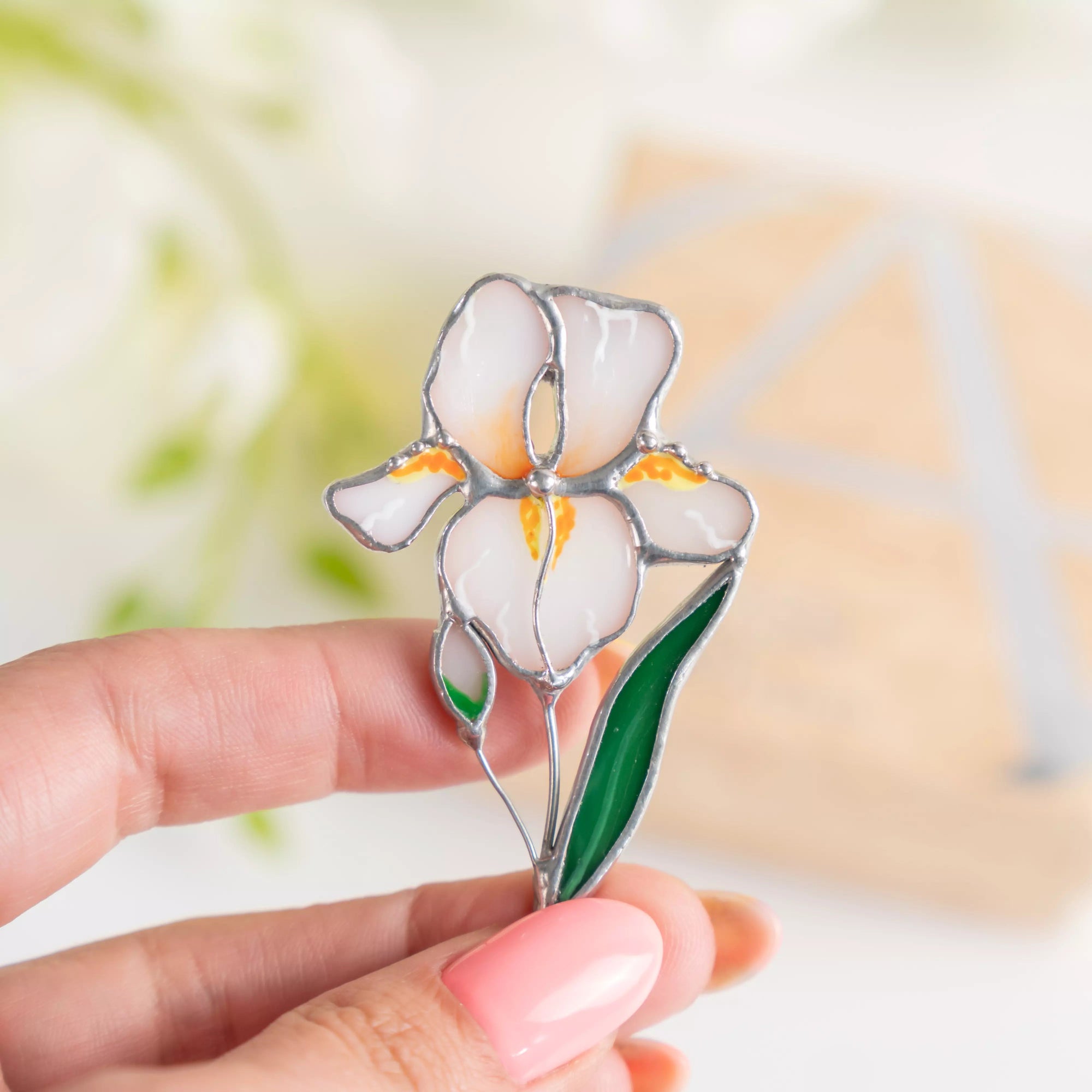 Hand holding a stained glass iris brooch with white petals, yellow accents, and green leaves.