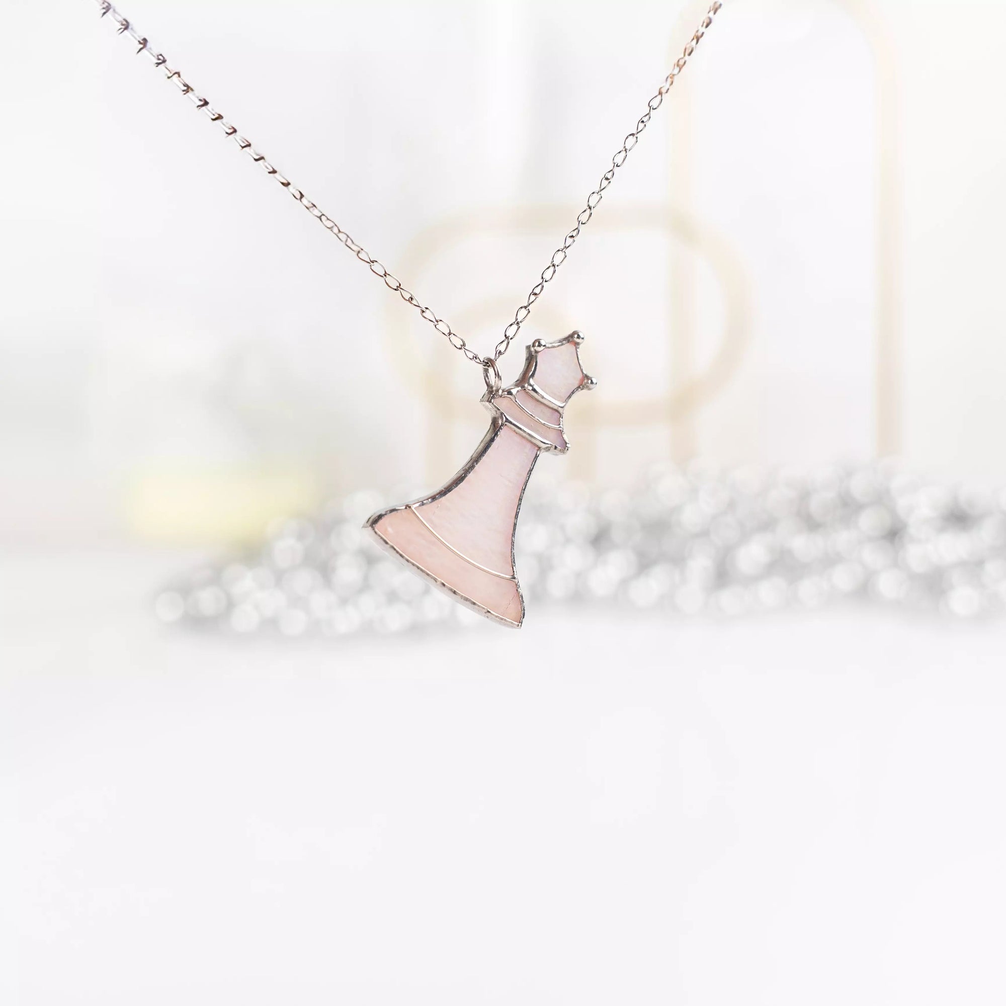 Light pink queen chess pendant hanging in air with blurred silver beads in background, soft and elegant atmosphere.