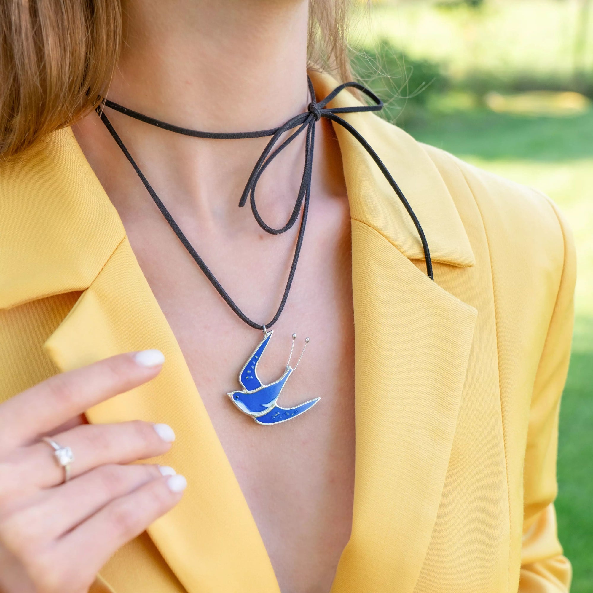 Blue stained glass swallow necklace styled with a mustard yellow blazer and tied black cord around the neck.