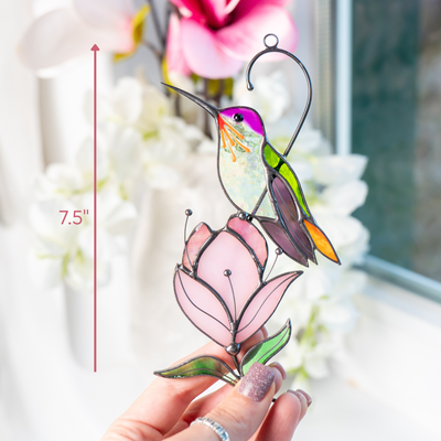 A hand holds a stained glass hummingbird sitting on a pink magnolia, showing the suncatcher size as 7.5 inches.