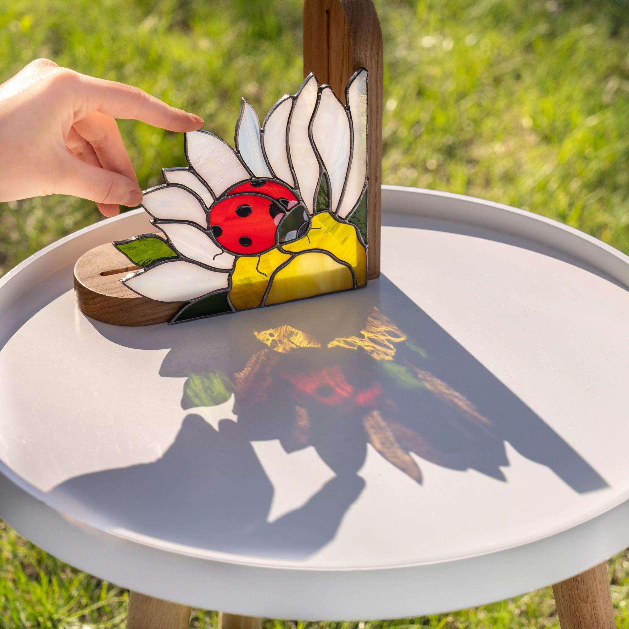 Stained glass daisy with a ladybug casting a beautiful colorful shadow on a white surface under the sun.