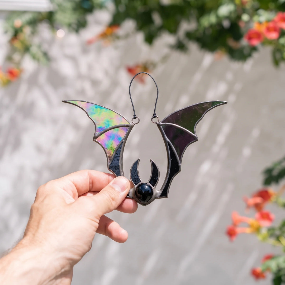 Iridescent bat Halloween stained glass suncatcher held against a sunlit white wall with red trumpet flowers behind - handmade gothic decor for a window or patio