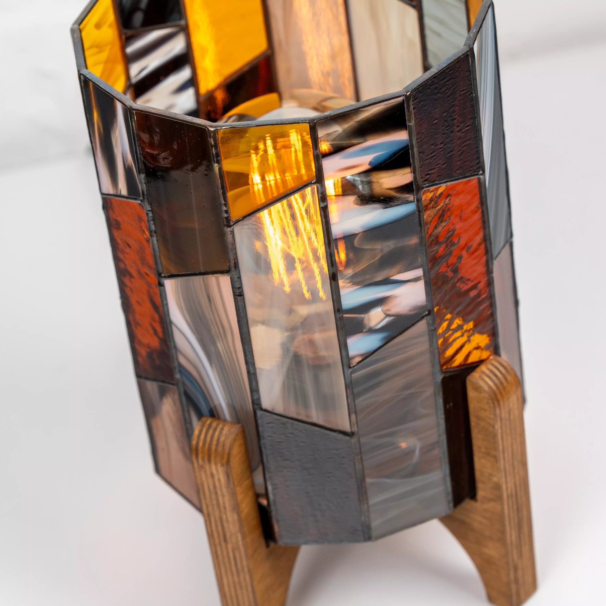 Geometric stained glass lamp in earthy amber and gray tones standing on wooden base, modern handmade nightstand light for cozy spaces.