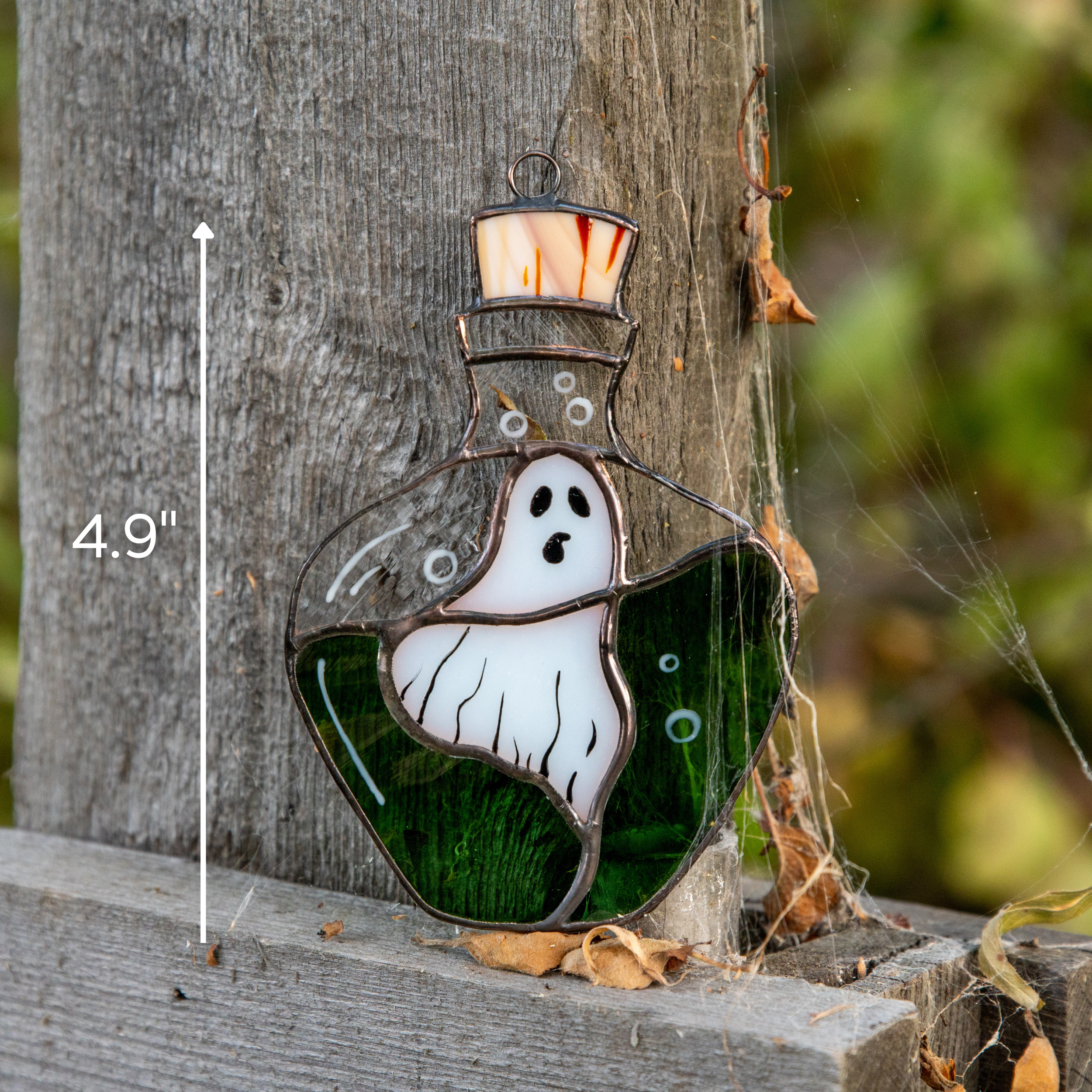 Stained glass ghost inside green potion bottle leaning against wood post outdoors with spider webs and size marking 4.9 inches.
