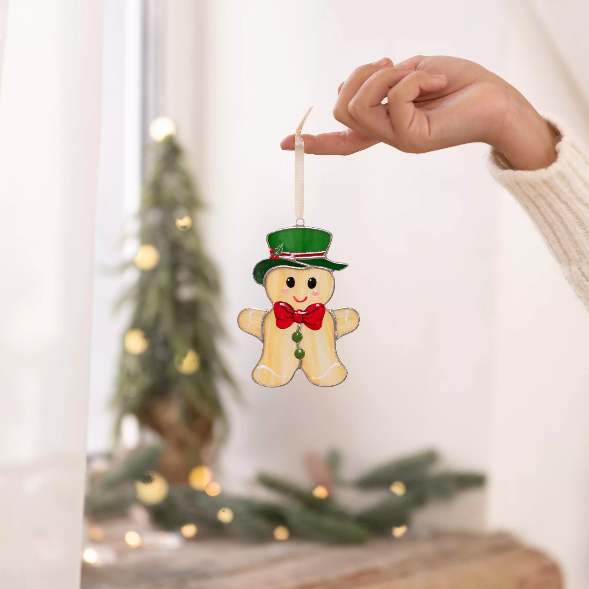 Hand holding a gingerbread man stained glass ornament with a green hat near a bright window.