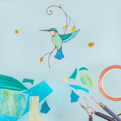 Stained glass hummingbird on a bright blue background, surrounded by colored glass shards, copper tape, and pliers