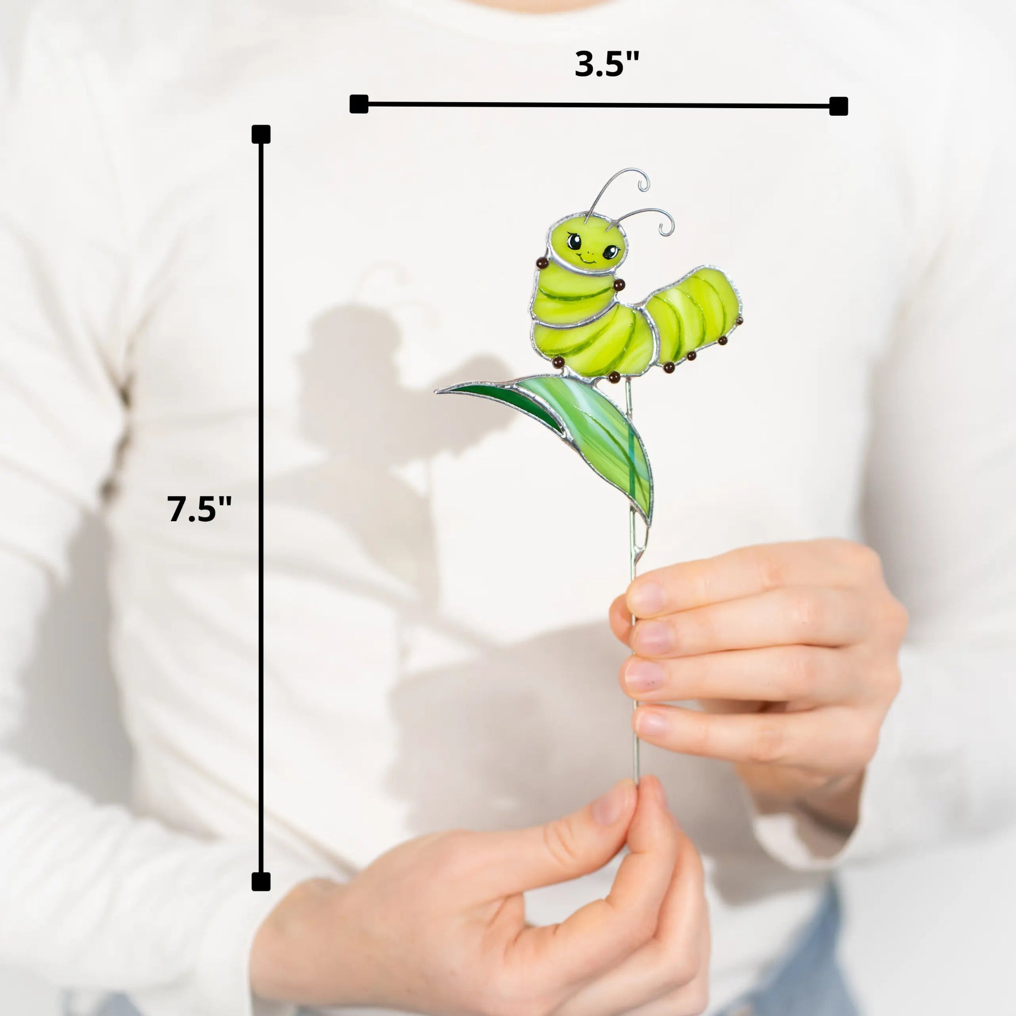 Stained glass caterpillar on a leaf with measurements 3.5 x 7.5 inches held by hands against a white background