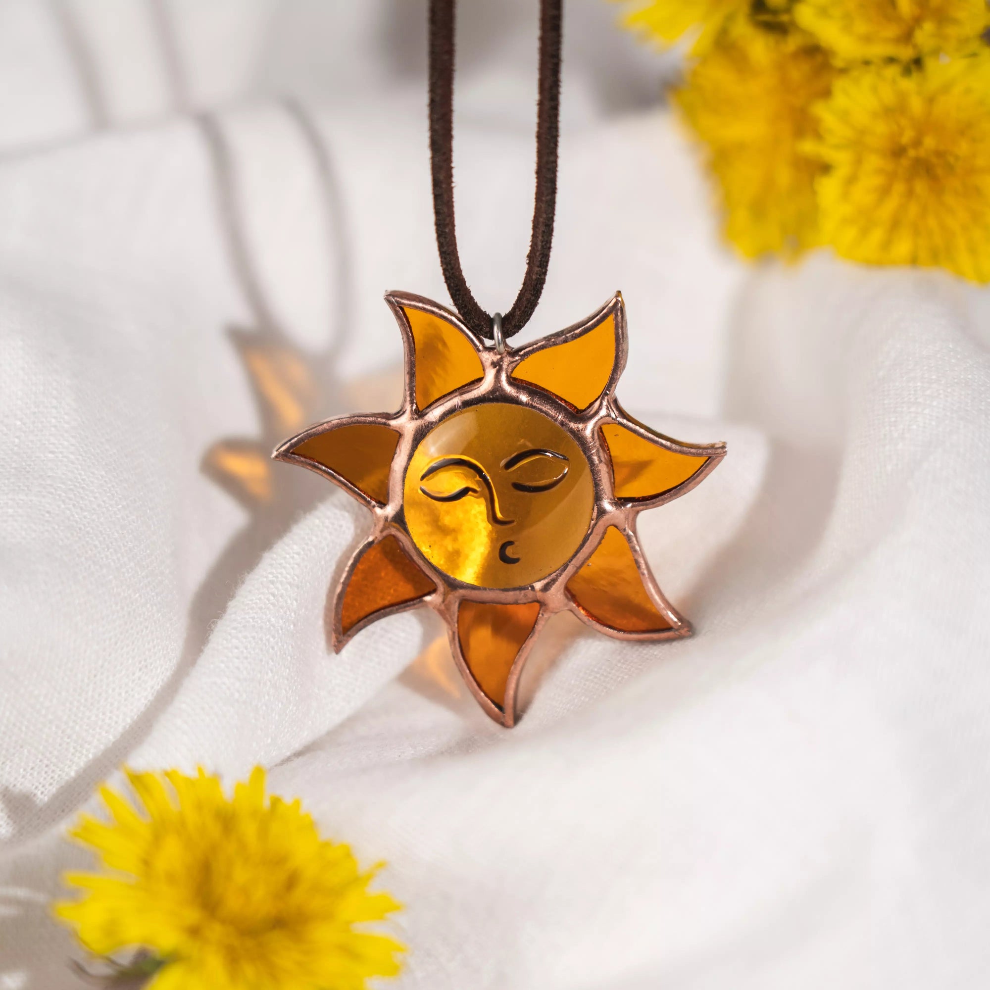 Amber stained glass sun necklace styled with yellow dandelions and white fabric background