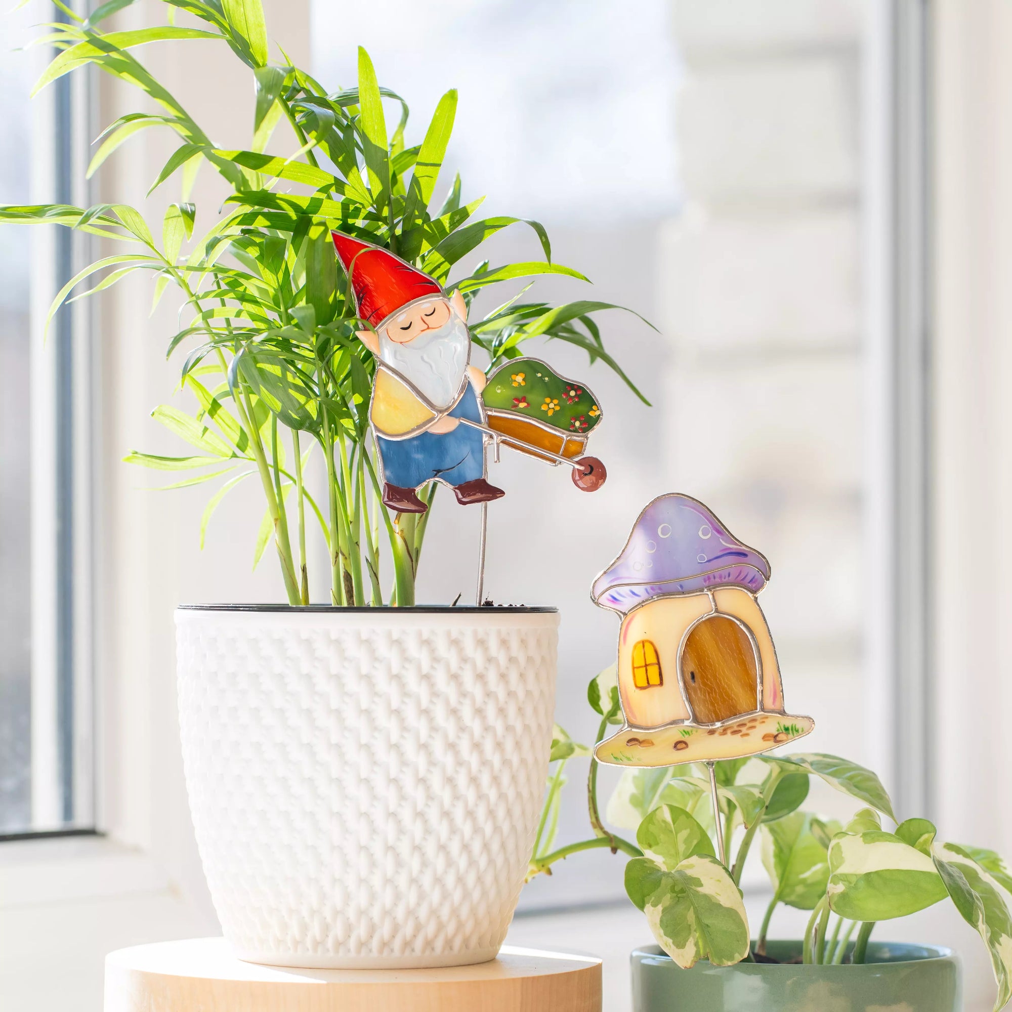 Stained glass gnome and mushroom house garden stakes on a plant with a blurred window background