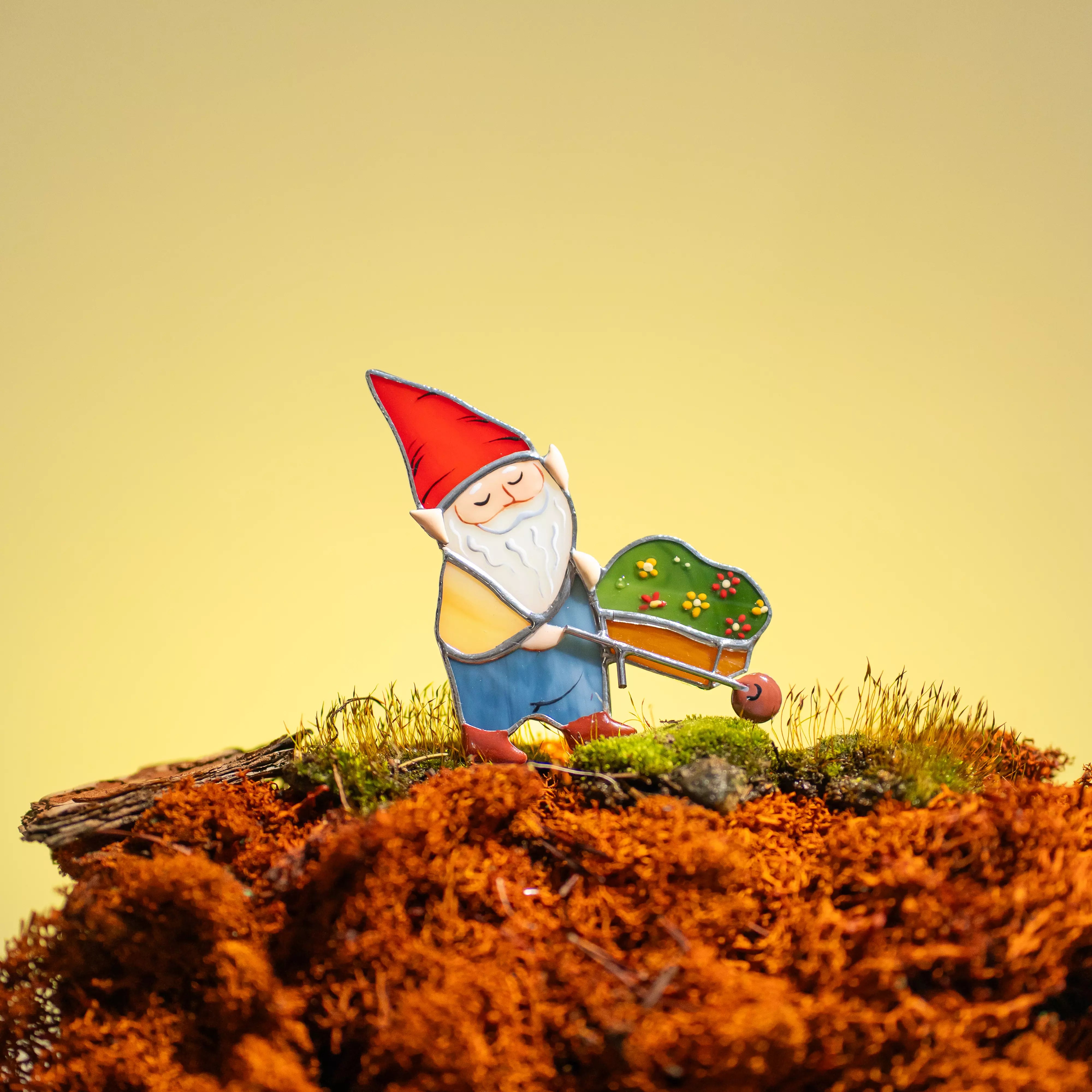 Stained glass plant stake nome with a red hat and white beard standing on a bed of red moss against a yellow background