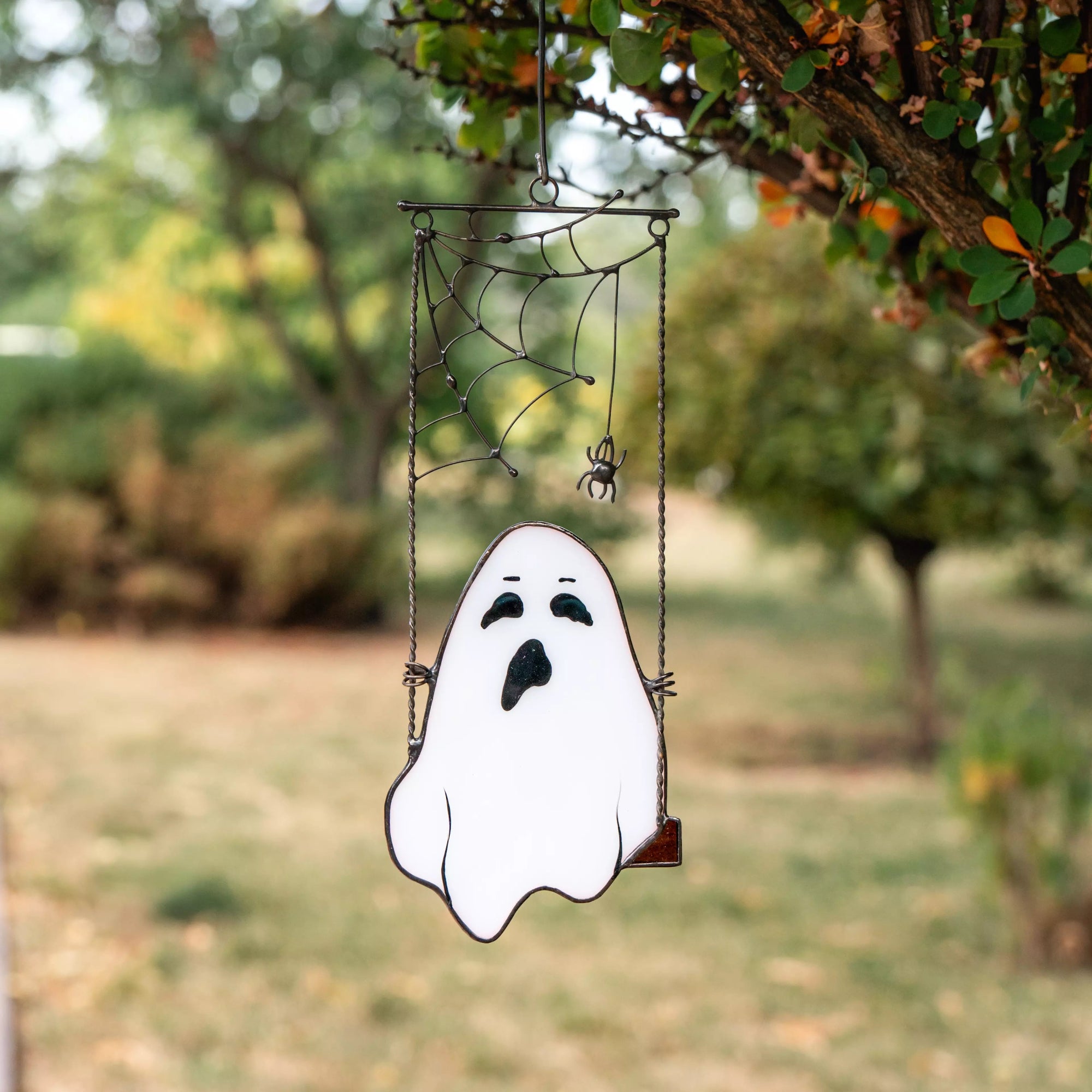 Ghost stained glass ornament with spiderweb hanging on tree branch outdoors