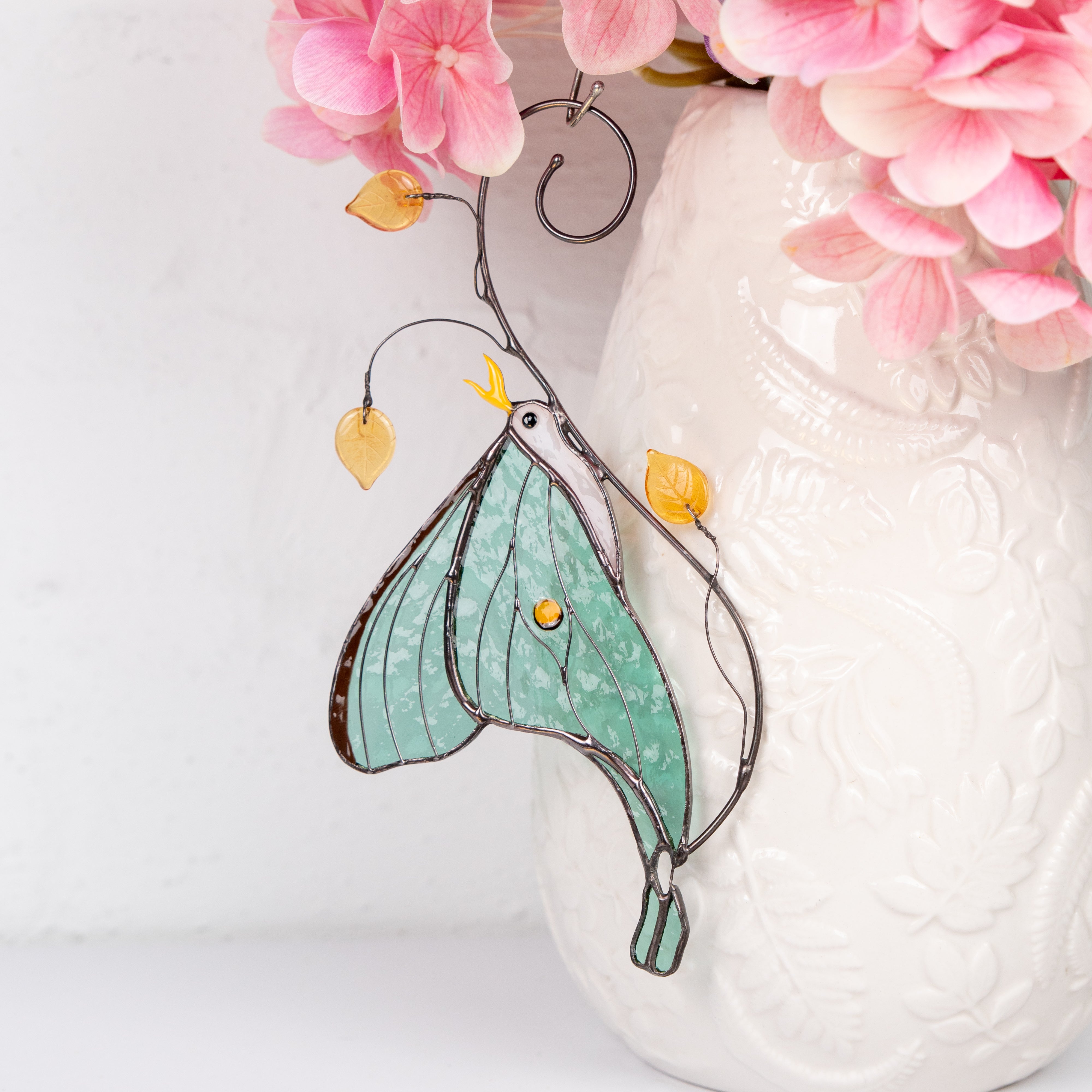 Luna moth stained glass suncatcher hanging against a white background with flowers nearby.