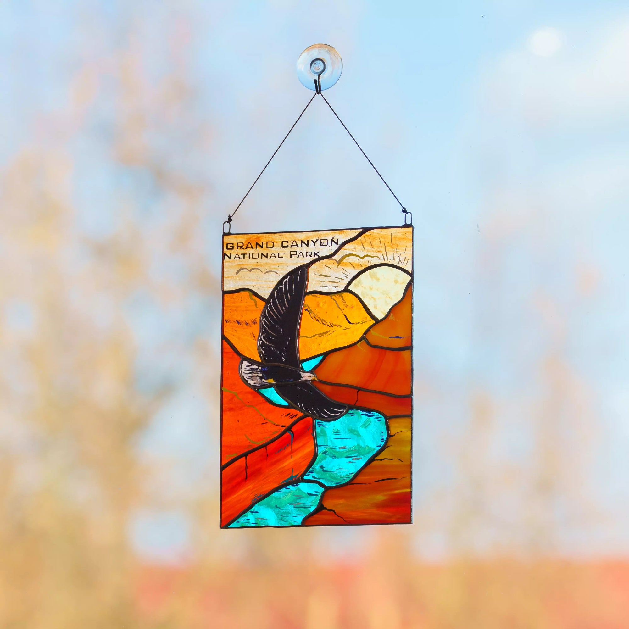 Stained glass panel of a bird over water with 'Grand Canyon National Park' text, hanging against a blurred natural background.