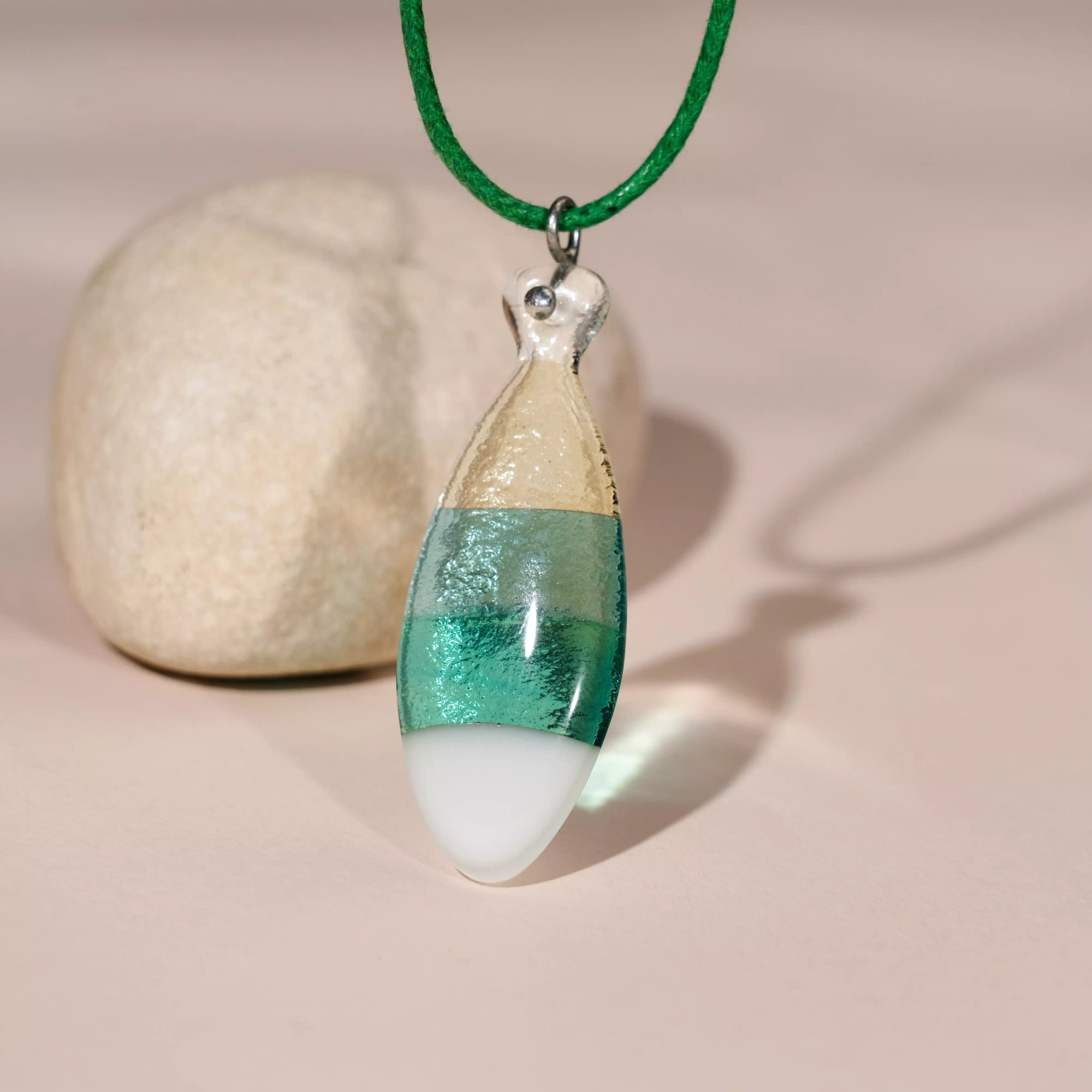 Sandy Green fused glass fish necklace with green cord displayed in front of a round beige stone on a soft pink surface.

