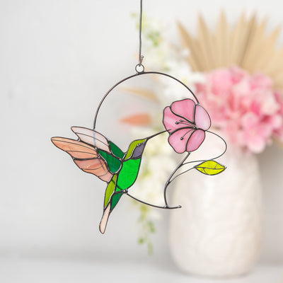 Colorful stained glass hummingbird with pink wings and flower, hanging in front of soft floral background